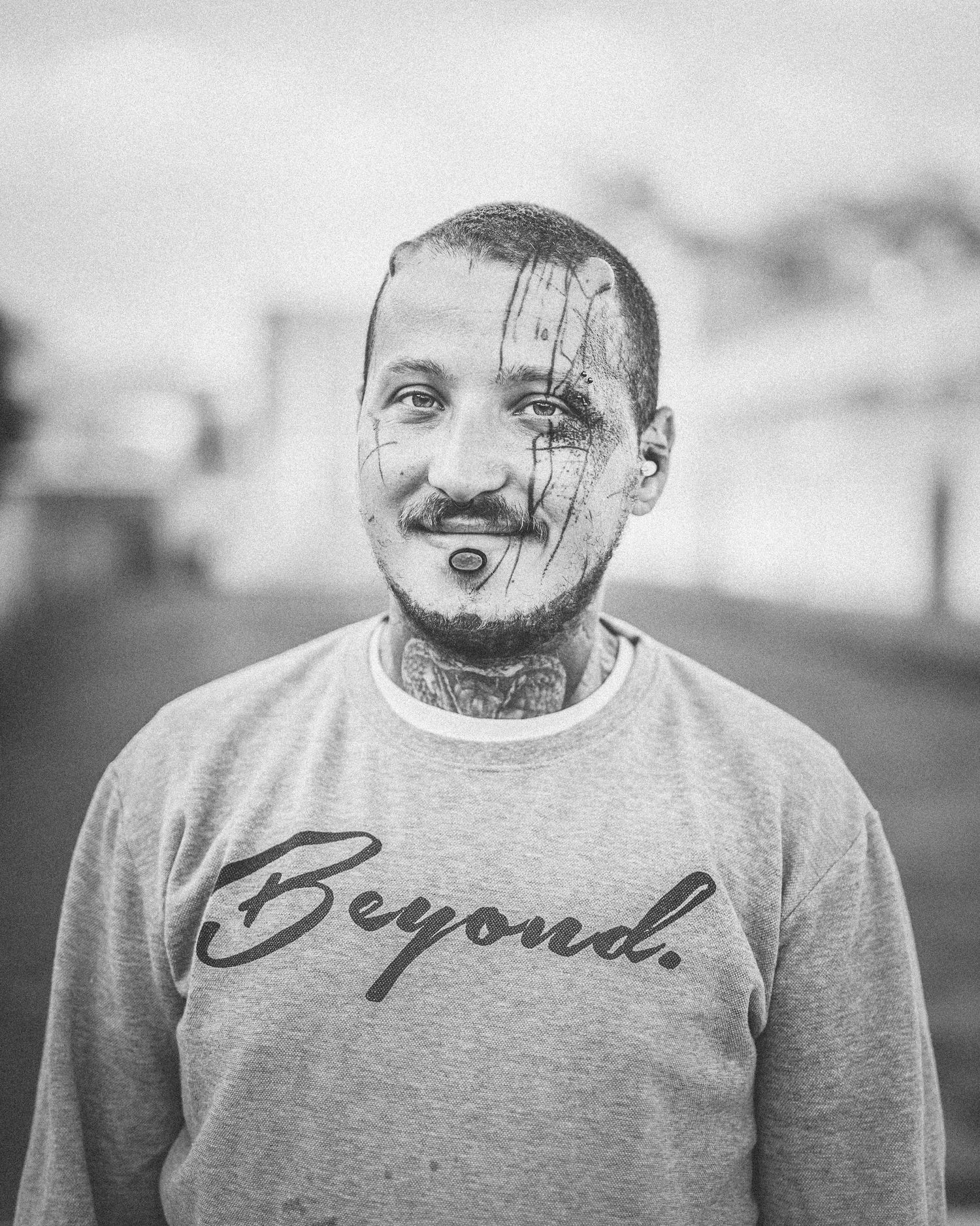 Black and white portrait of a tattooed man with facial ink smears wearing a gray Beyond sweatshirt outdoors.