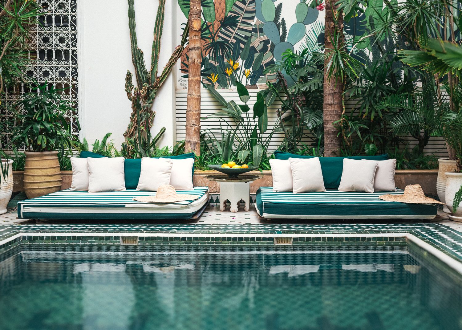 Luxury pool area with teal daybeds, white cushions, lush tropical plants, and a decorative mosaic tile surround.
