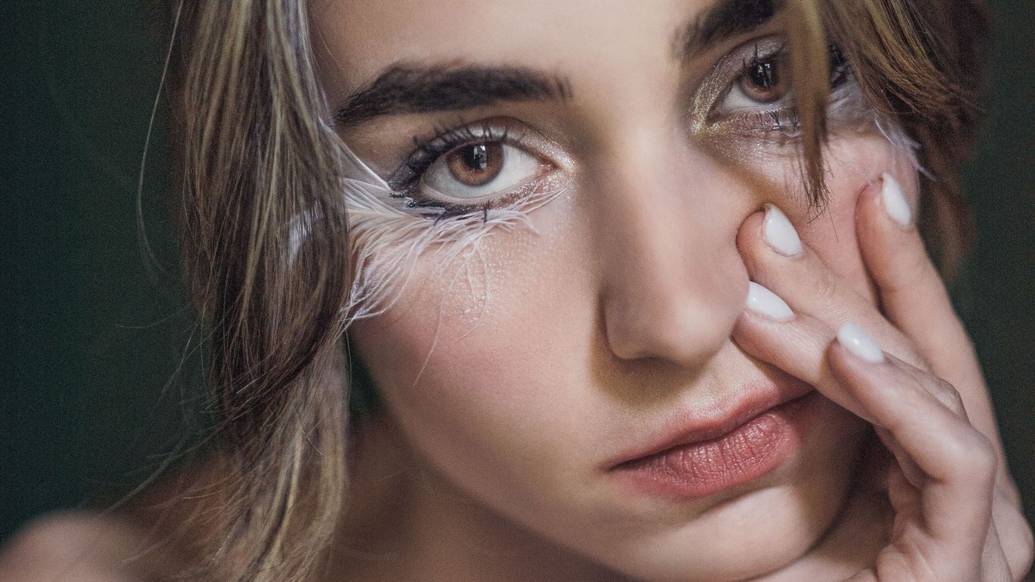 Close-up portrait of a young woman with artistic white feather eye makeup touching her face gently.