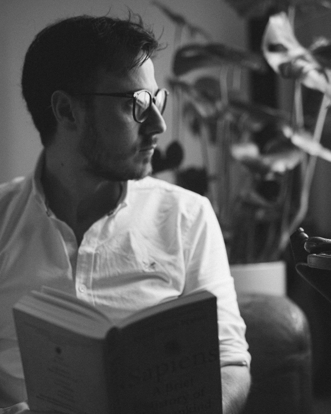 Black and white photo of a bearded man wearing glasses reading a book in a cozy indoor setting.