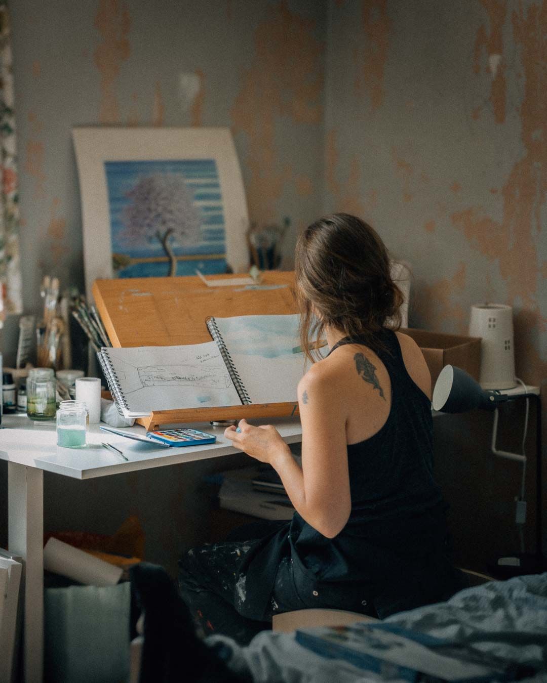Woman with tattoo sits at art desk sketching in sketchbook, with painting and art supplies in rustic studio.