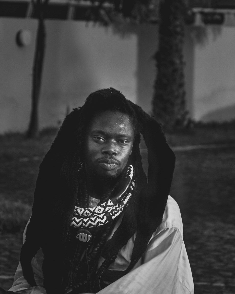 Black and white portrait of a man with dreadlocks and beaded necklace seated outdoors near a palm tree.