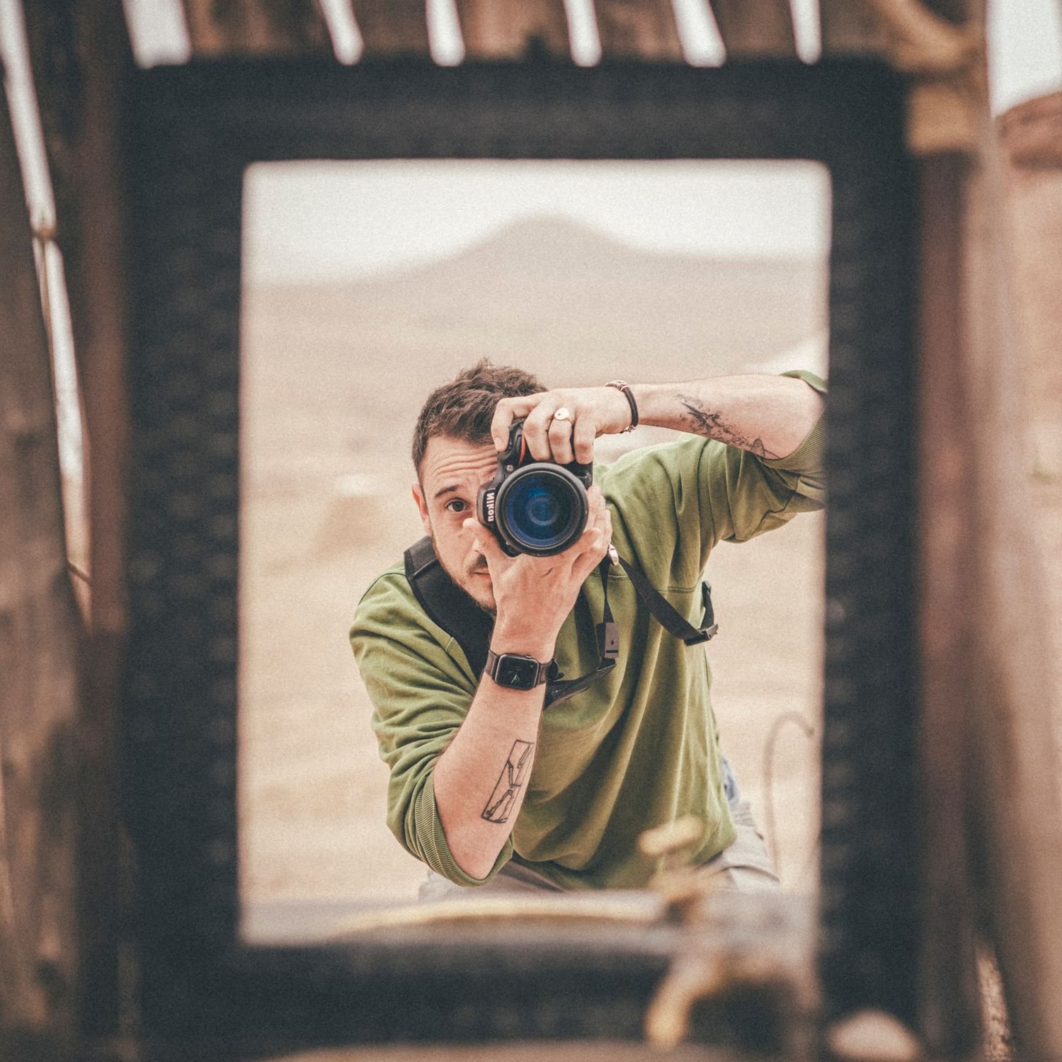 Photographer capturing a self-portrait through a rustic wooden frame with arid landscape in the background.