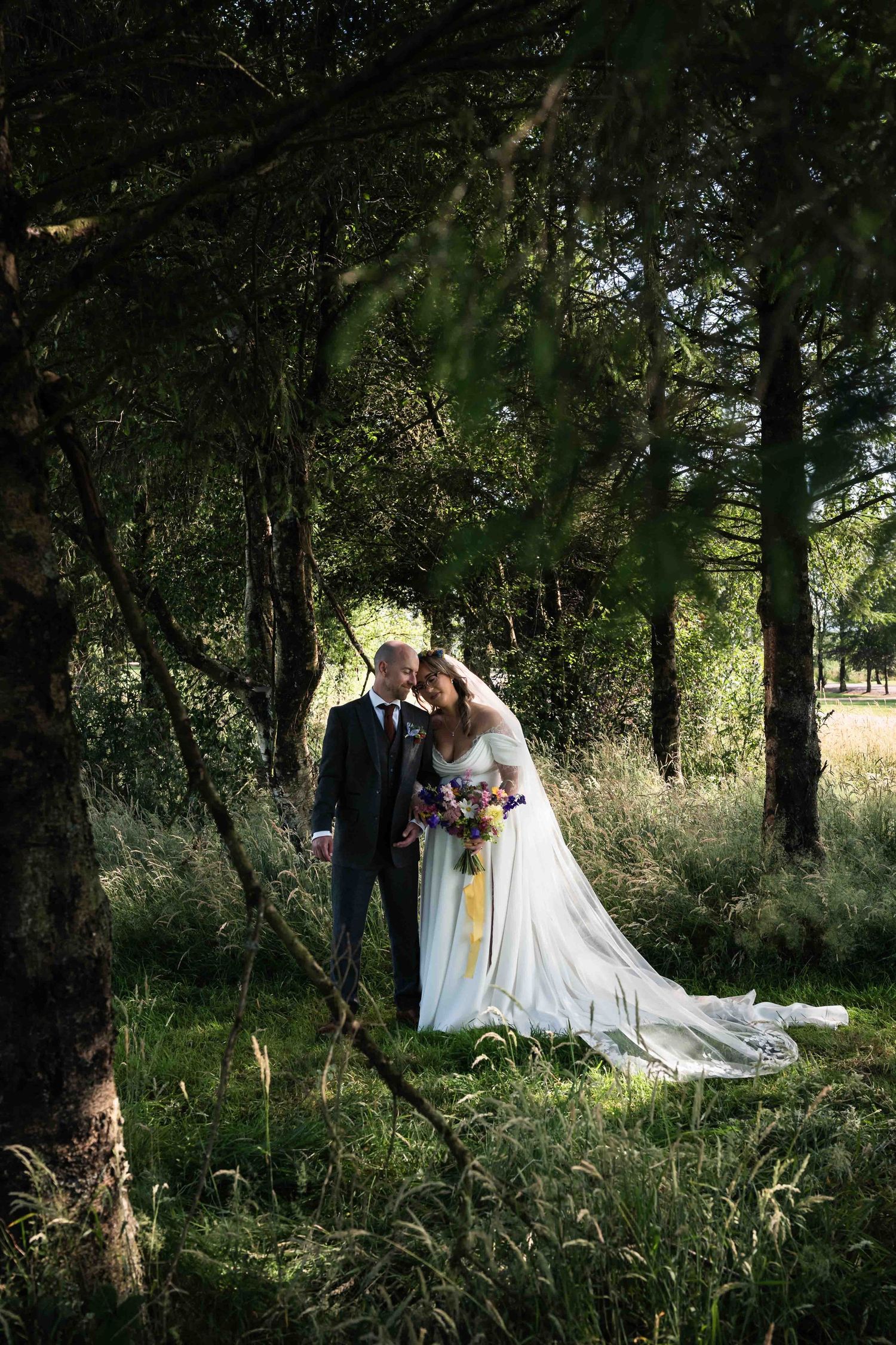Bride and groom embrace romantically in a lush green forest clearing, bride holding a colorful bouquet. Mountain View, Kilkenny