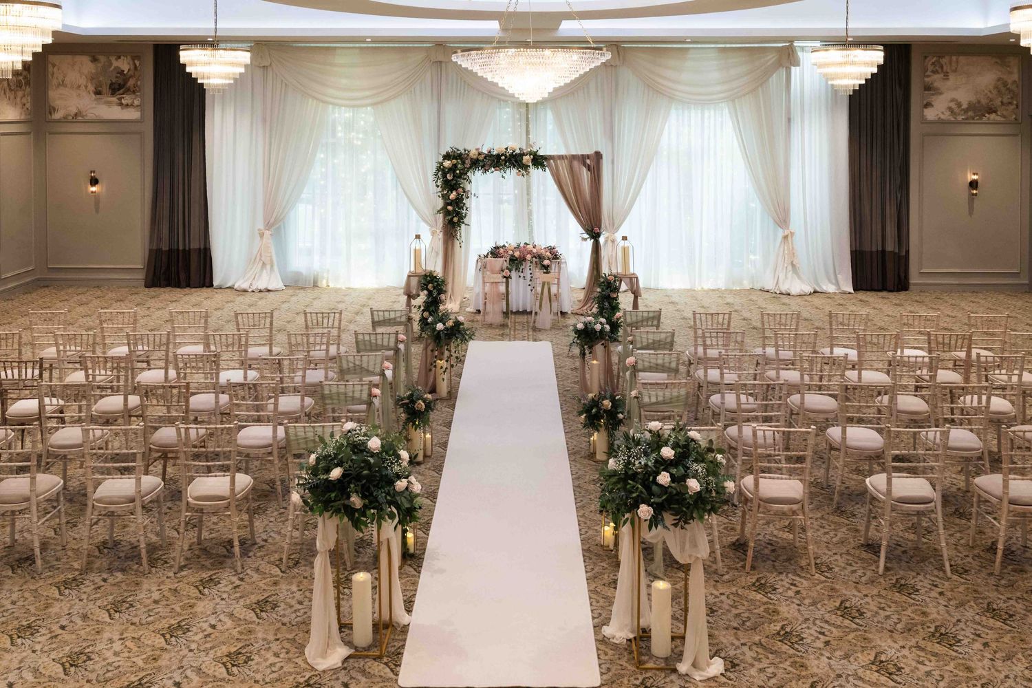 Elegant wedding ceremony setup with white aisle runner, floral arch, candles, and chiavari chairs in a grand ballroom. Dunboyne Castle