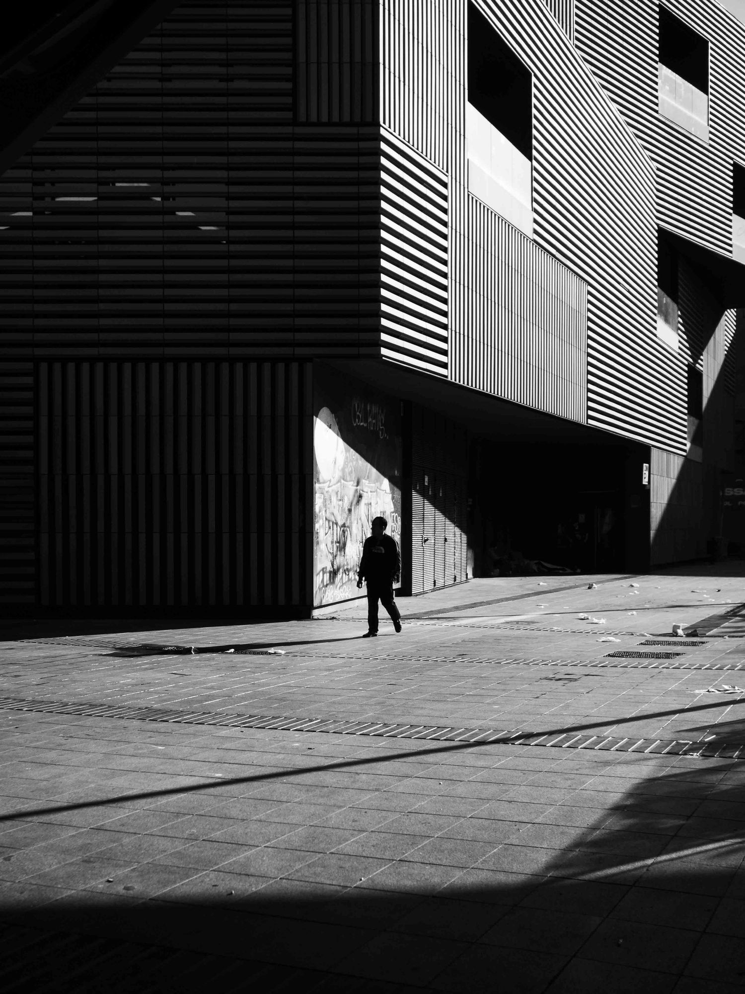 Lone pedestrian silhouette walking past modern striped building facade with dramatic shadows in black and white.