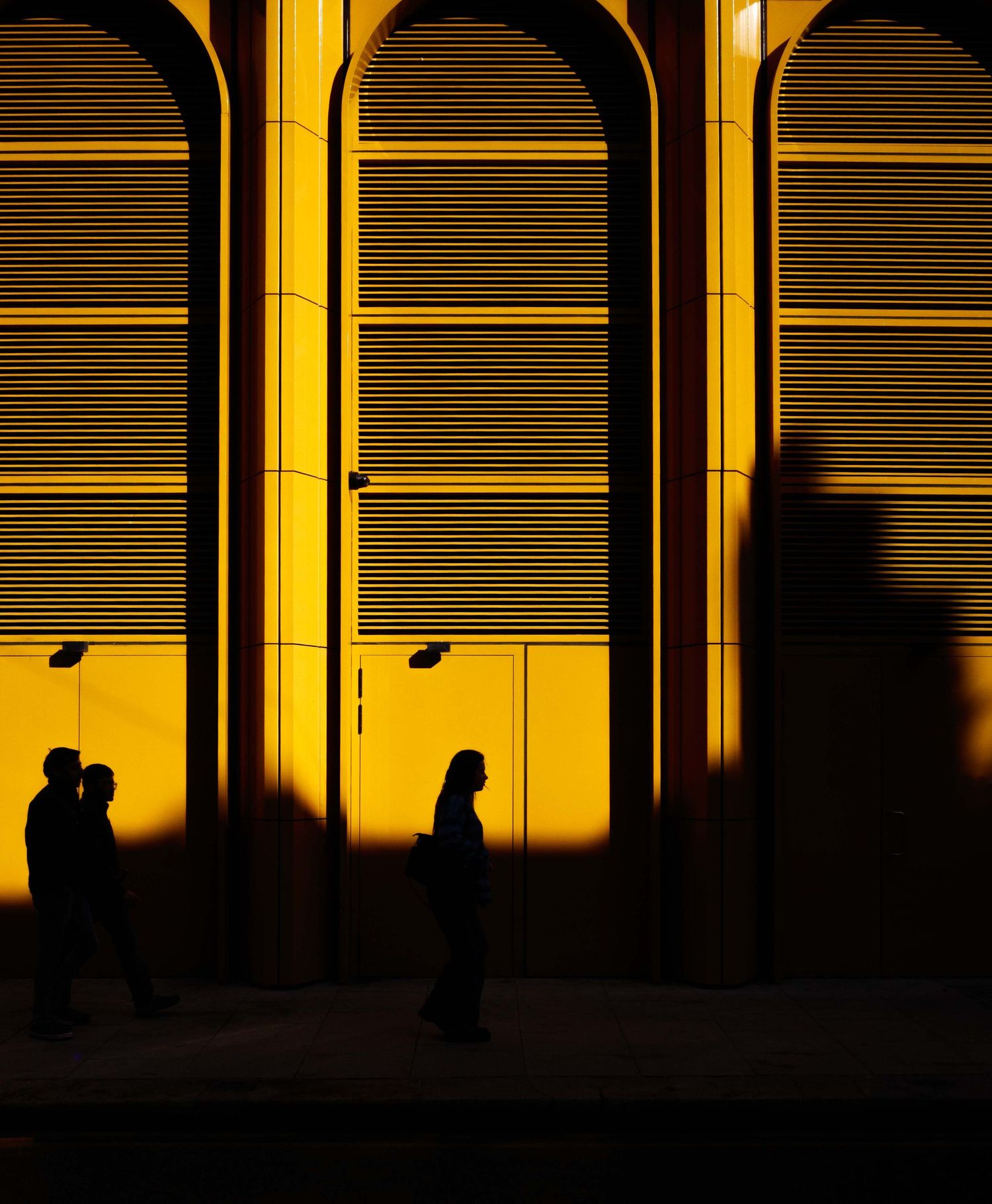 Silhouettes of pedestrians walk past a golden yellow arched building facade at sunset.