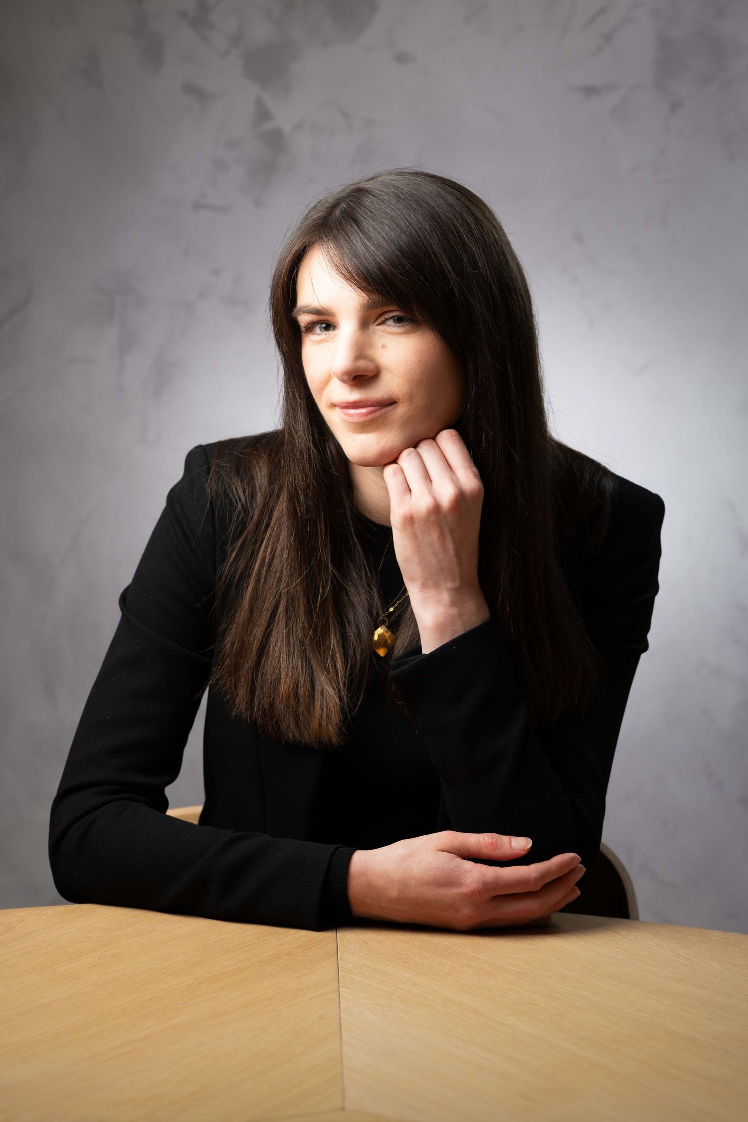 Young woman with long dark hair in black blazer sitting at wooden table resting chin on hand.  Headshot Photography Ireland