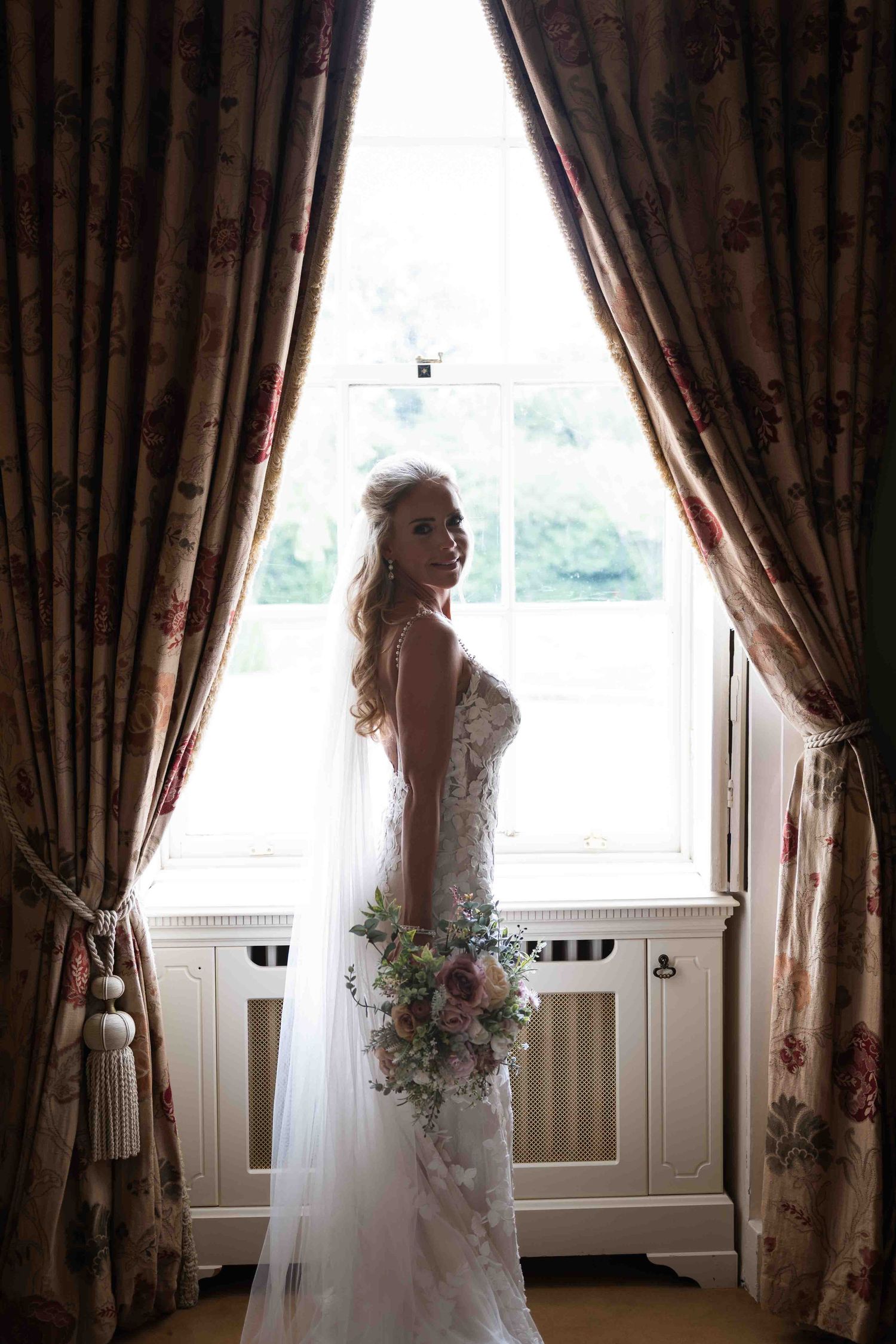 Elegant bride in a lace gown holding a bouquet, standing before a large window with floral curtains. Dunboyne Castle