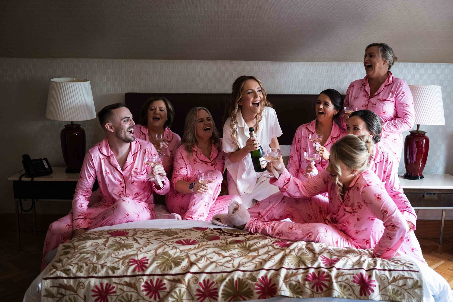 Bridal party laughing and celebrating in pink pajamas on a bed with champagne. Irish wedding photography. Langtons, Kilkenny