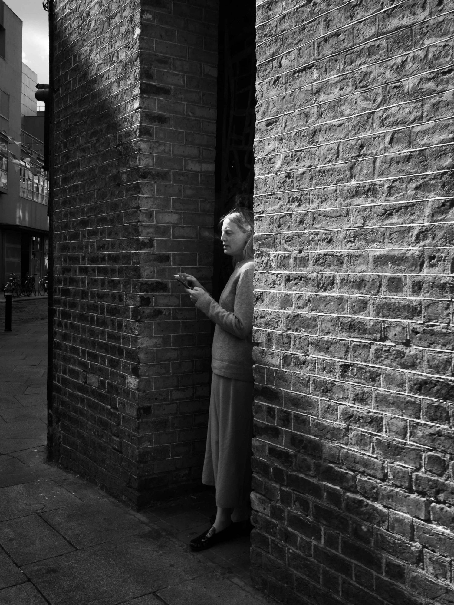 Black and white street photo of an elderly woman peering around a brick wall corner in an urban alley.