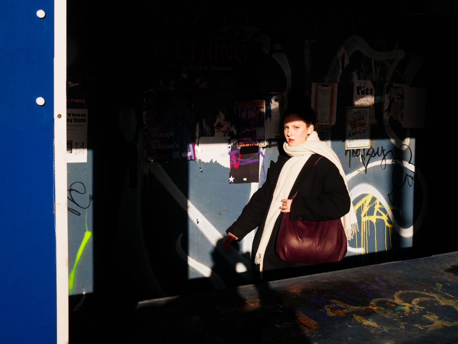 A woman carrying a burgundy bag walks through a graffiti-covered urban underpass in sunlight.