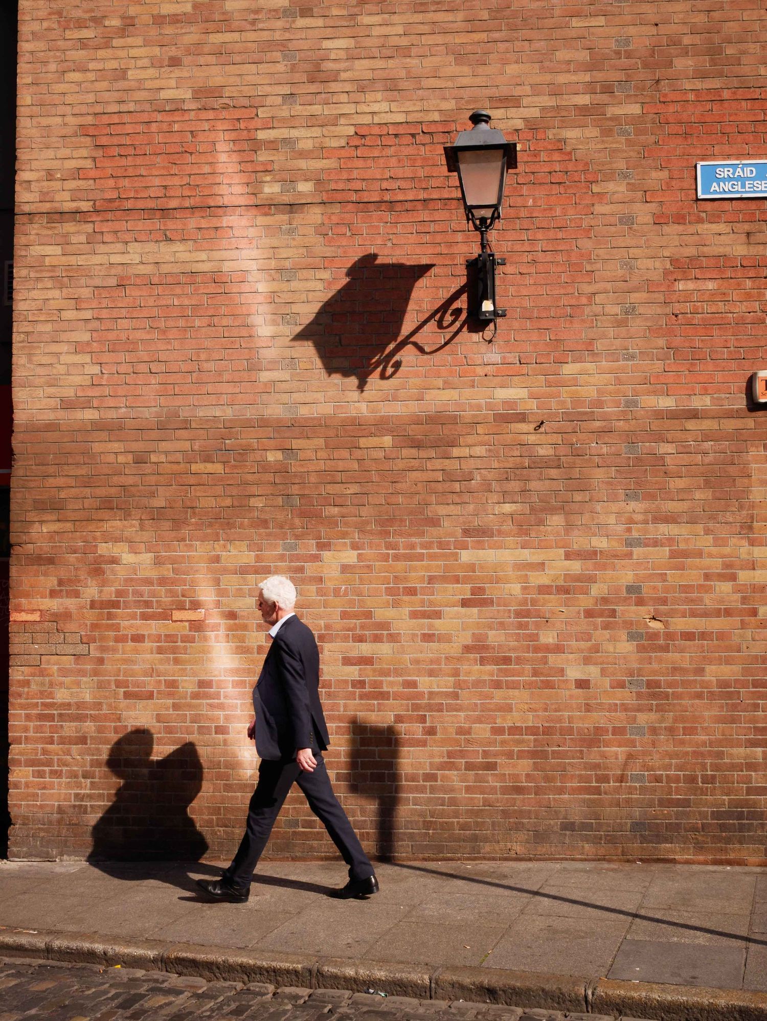Man in a suit walking past a red brick wall with a decorative street lamp casting shadows in sunlight.