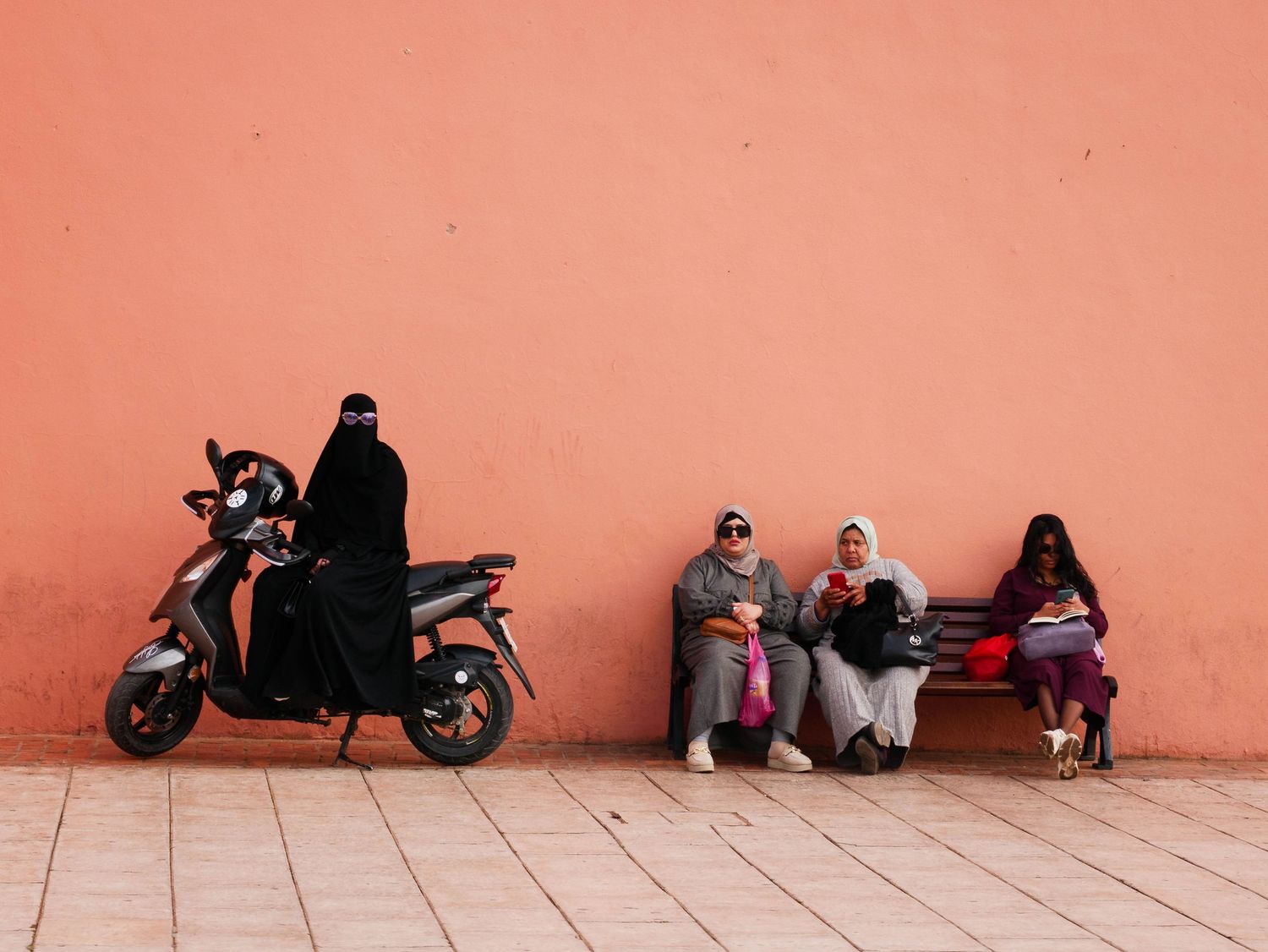 Three women sit on a bench beside a pink wall while another rides a moped in Morocco.