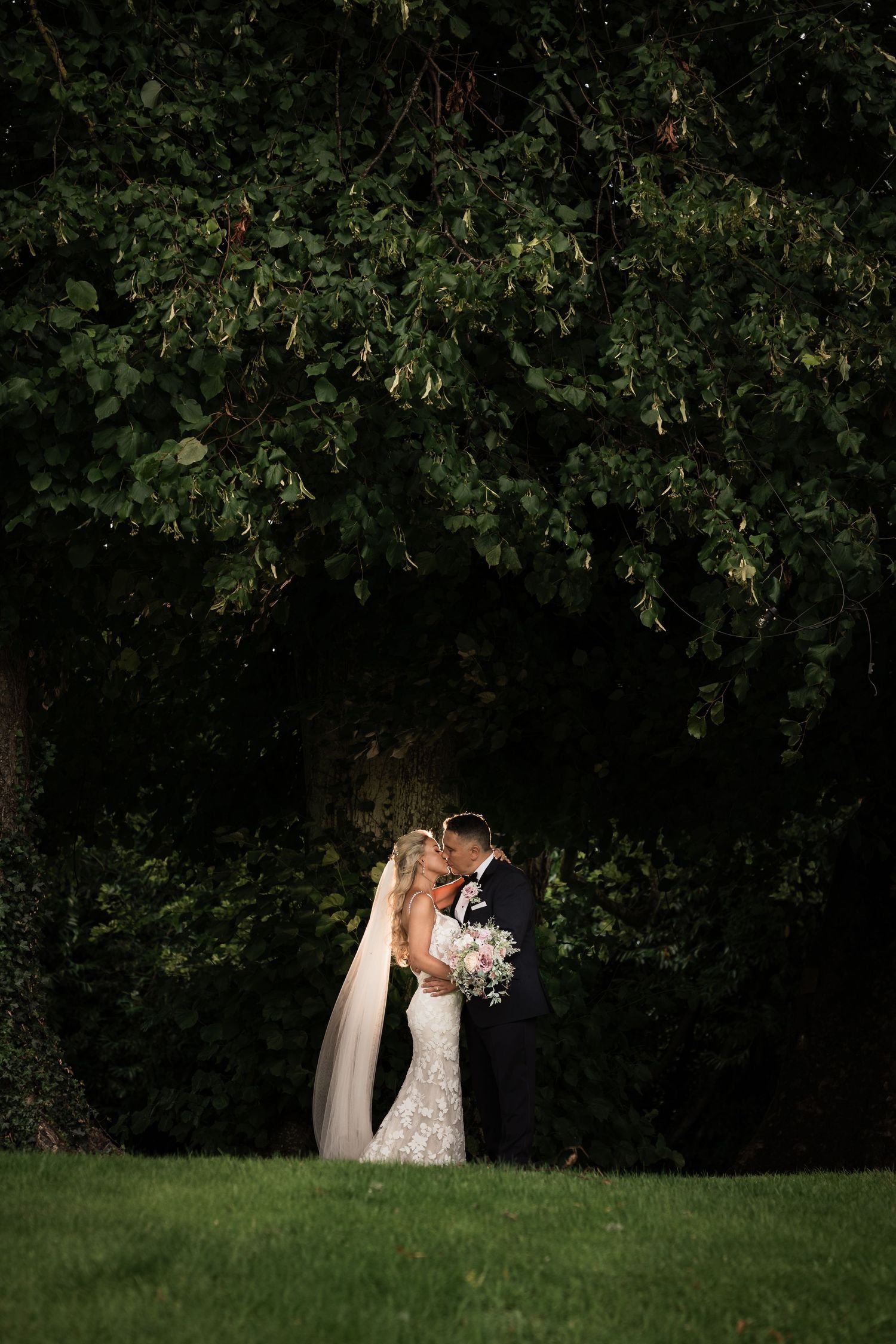 Bride and groom sharing a romantic kiss beneath a large tree in a lush green garden at dusk. Dunboyne Castle