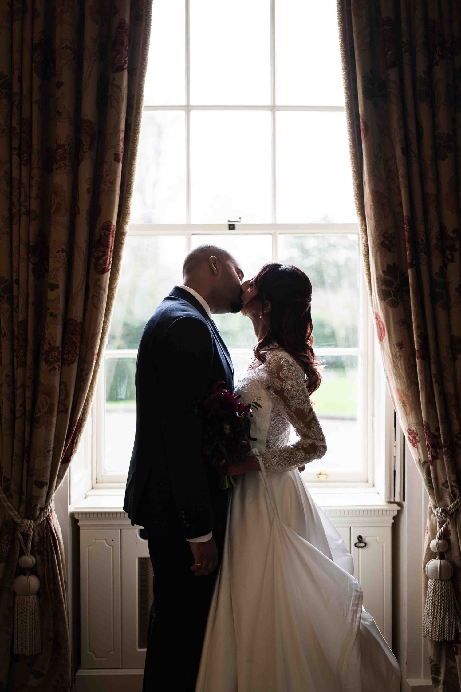 Bride and groom sharing a kiss in silhouette before a large window with floral curtains. Dunboyne Castle