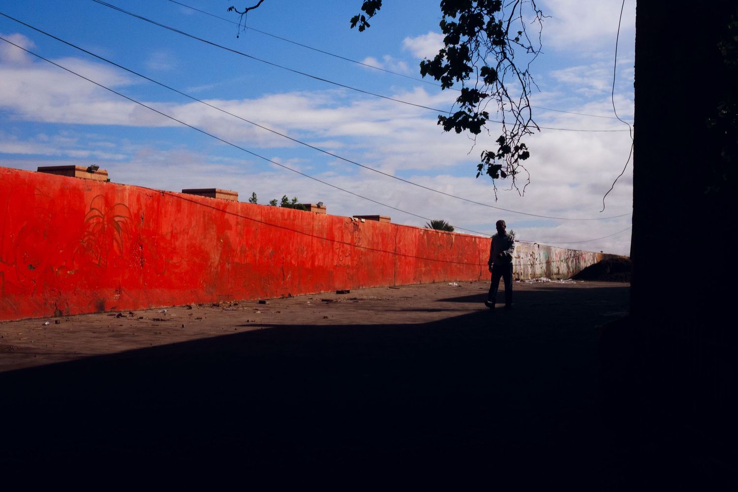A lone silhouetted figure stands beside a vibrant red painted wall under a blue sky.