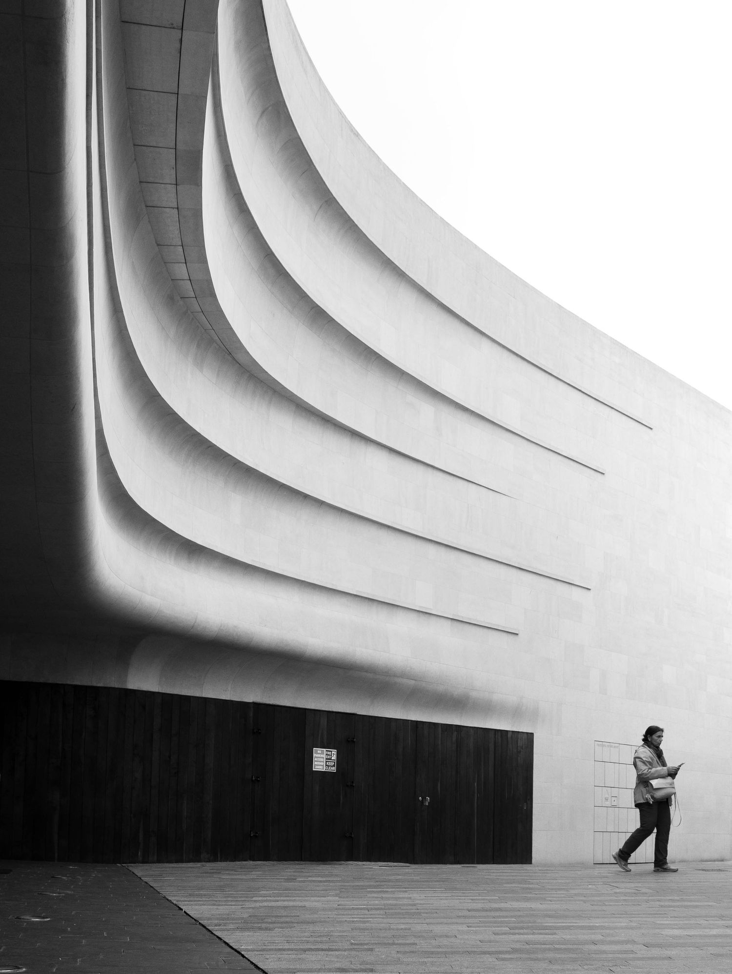 Black and white photo of a person walking past a modern curved concrete building facade.