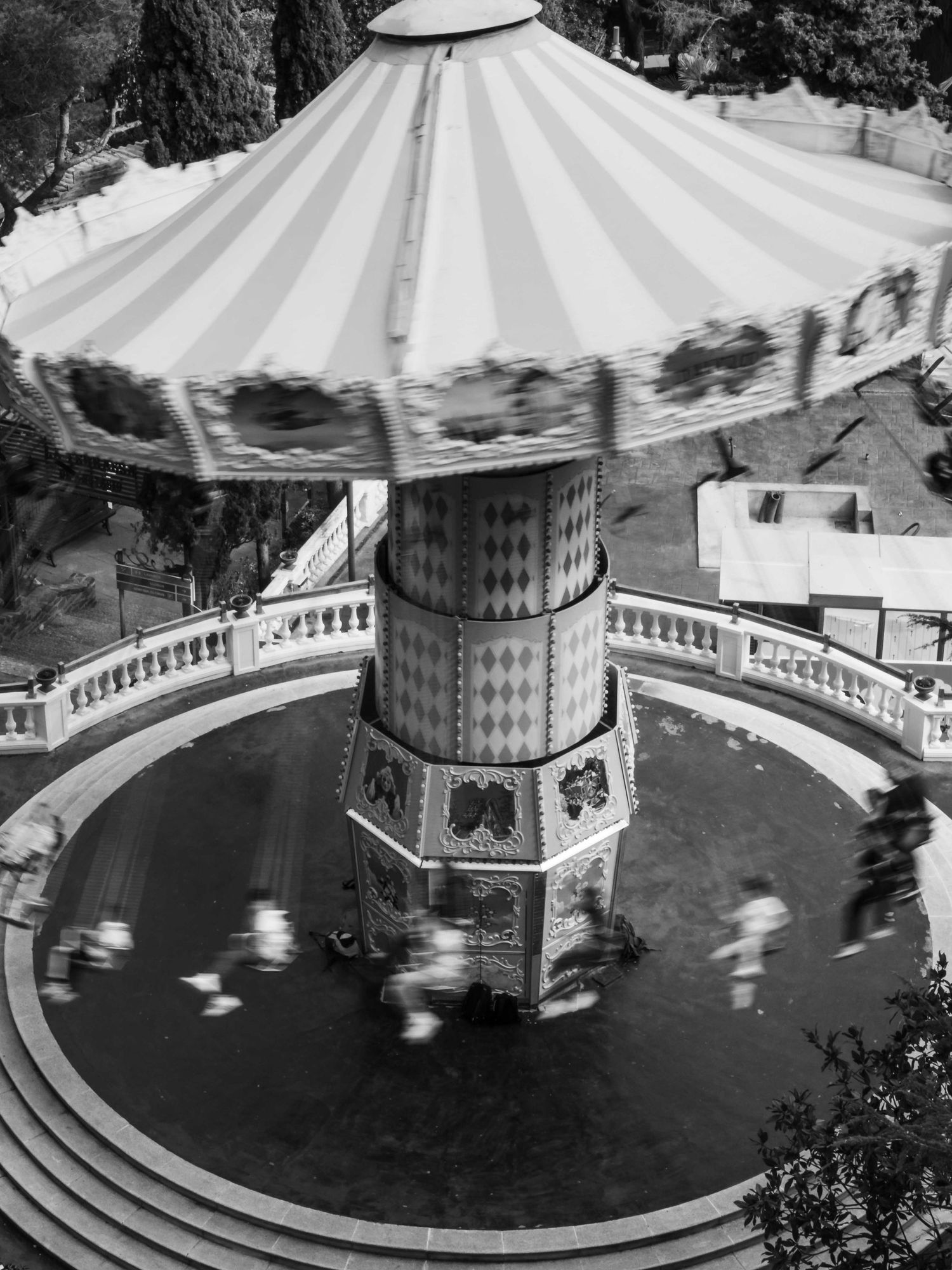 Aerial black and white long exposure shot of spinning carousel with blurred riders at amusement park.
