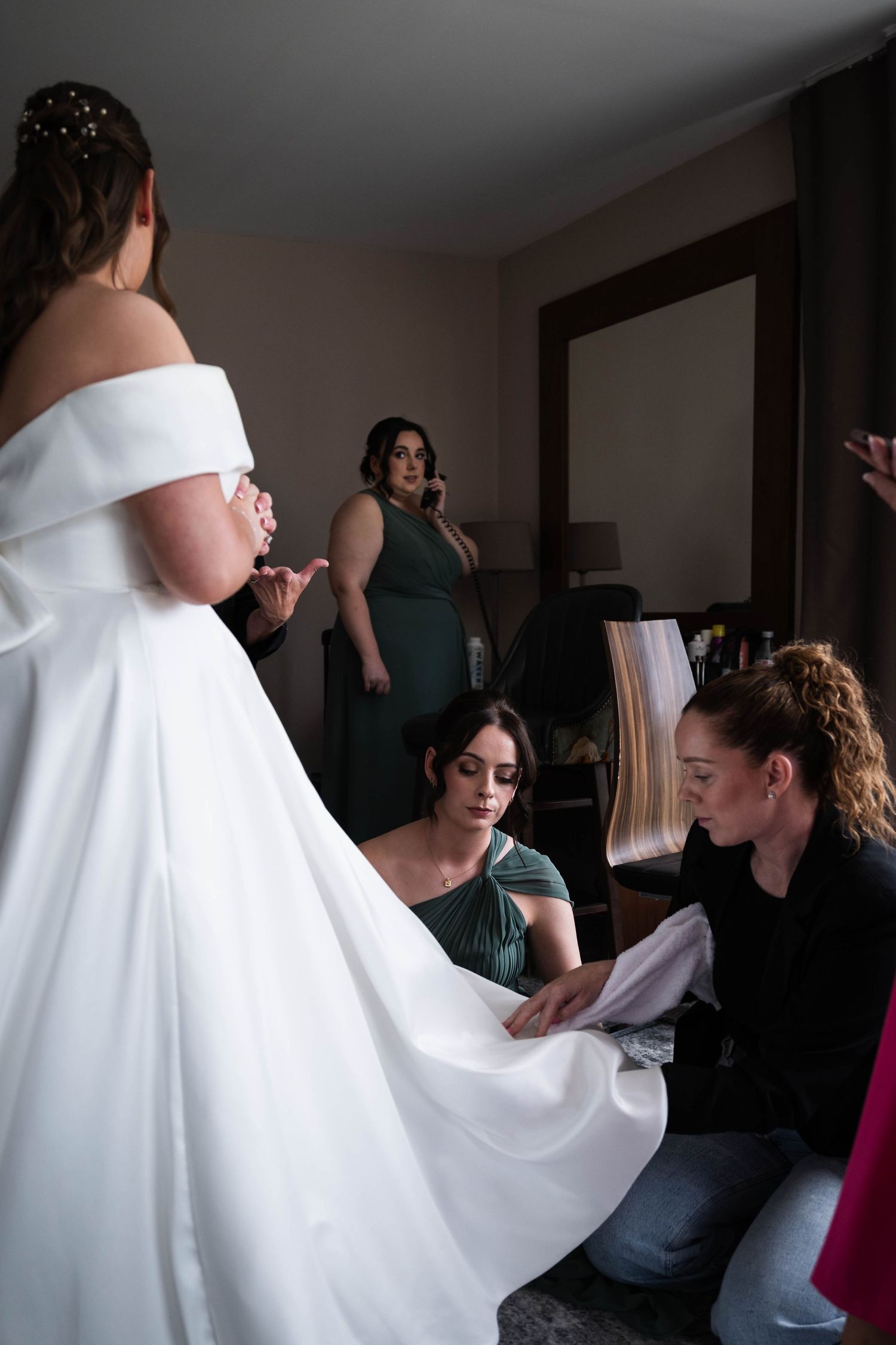 Bridesmaids and stylist carefully adjust the train of a brides white off-shoulder gown indoors. Irish wedding photography. Dunboyne Castle