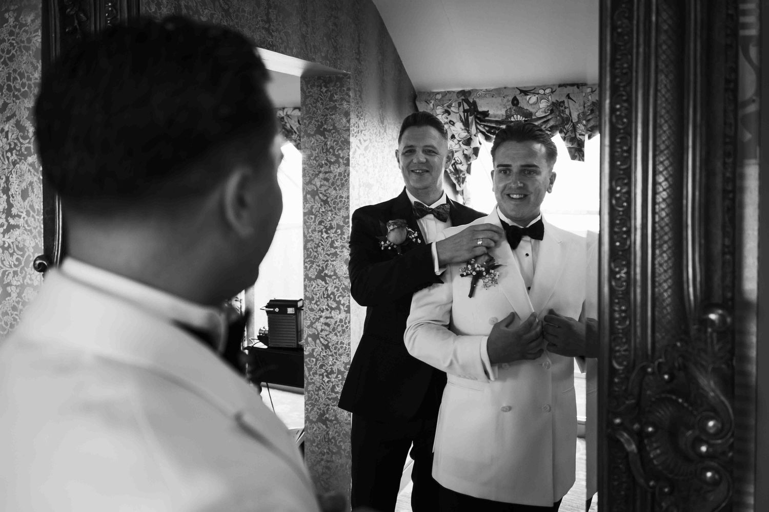 Two groomsmen in tuxedos prepare together in front of an ornate mirror in black and white photo. Queer wedding. Clontarf Castle