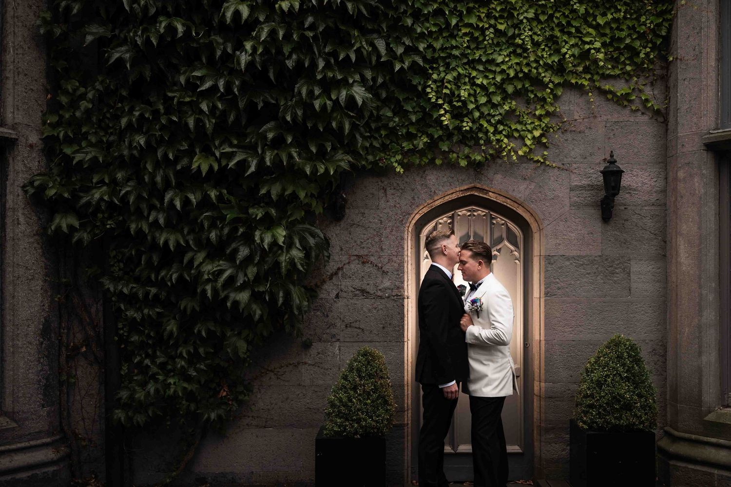 Two grooms share romantic moment in doorway surrounded by lush ivy-covered stone wall at wedding. Queer wedding. Clontarf Castle