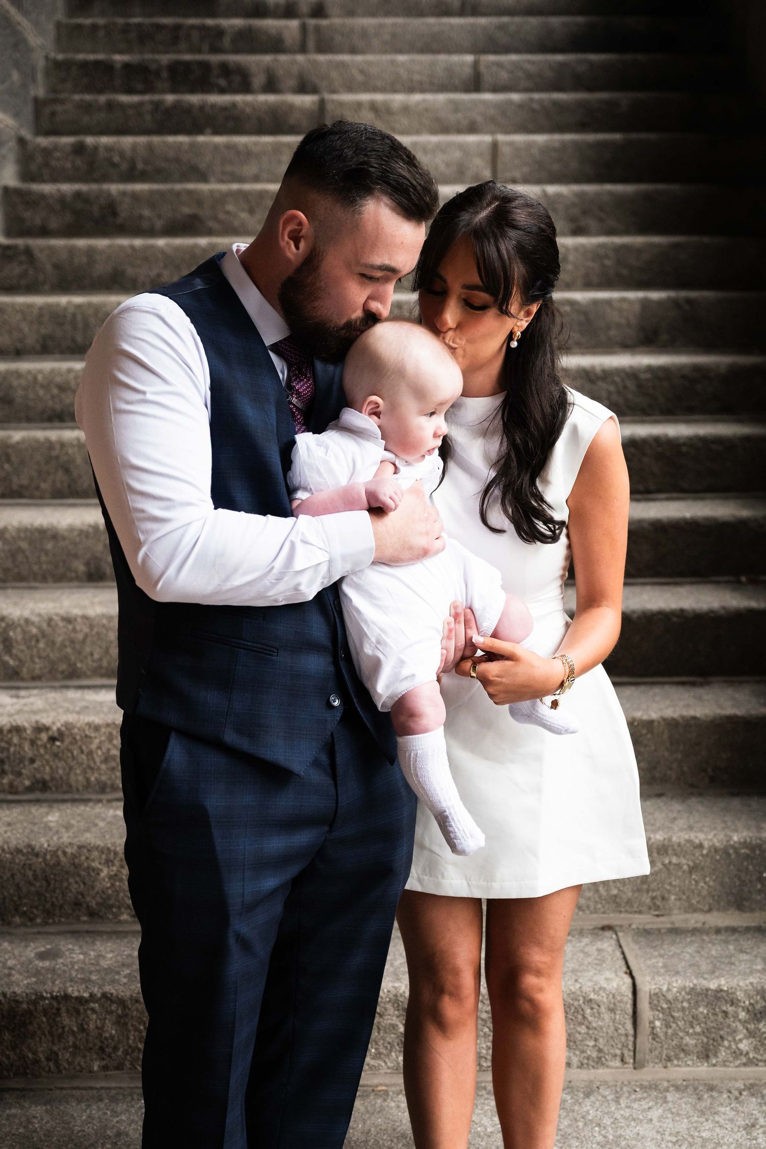 Couple in formal wear tenderly kissing their baby on stone steps. Christening Photography Ireland