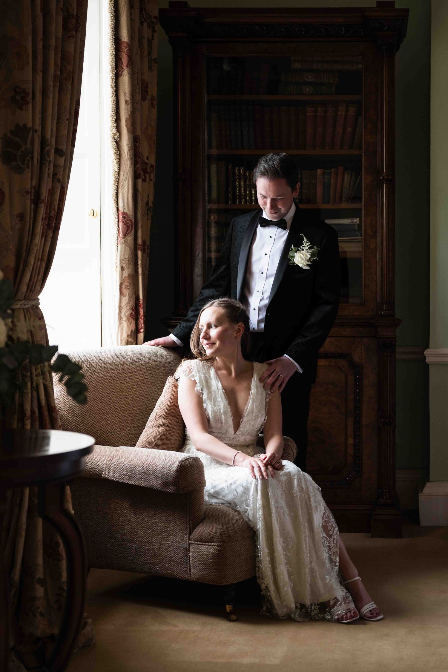 Bride in lace gown seated in armchair with groom in tuxedo standing beside her in a library. Dunboyne Castle