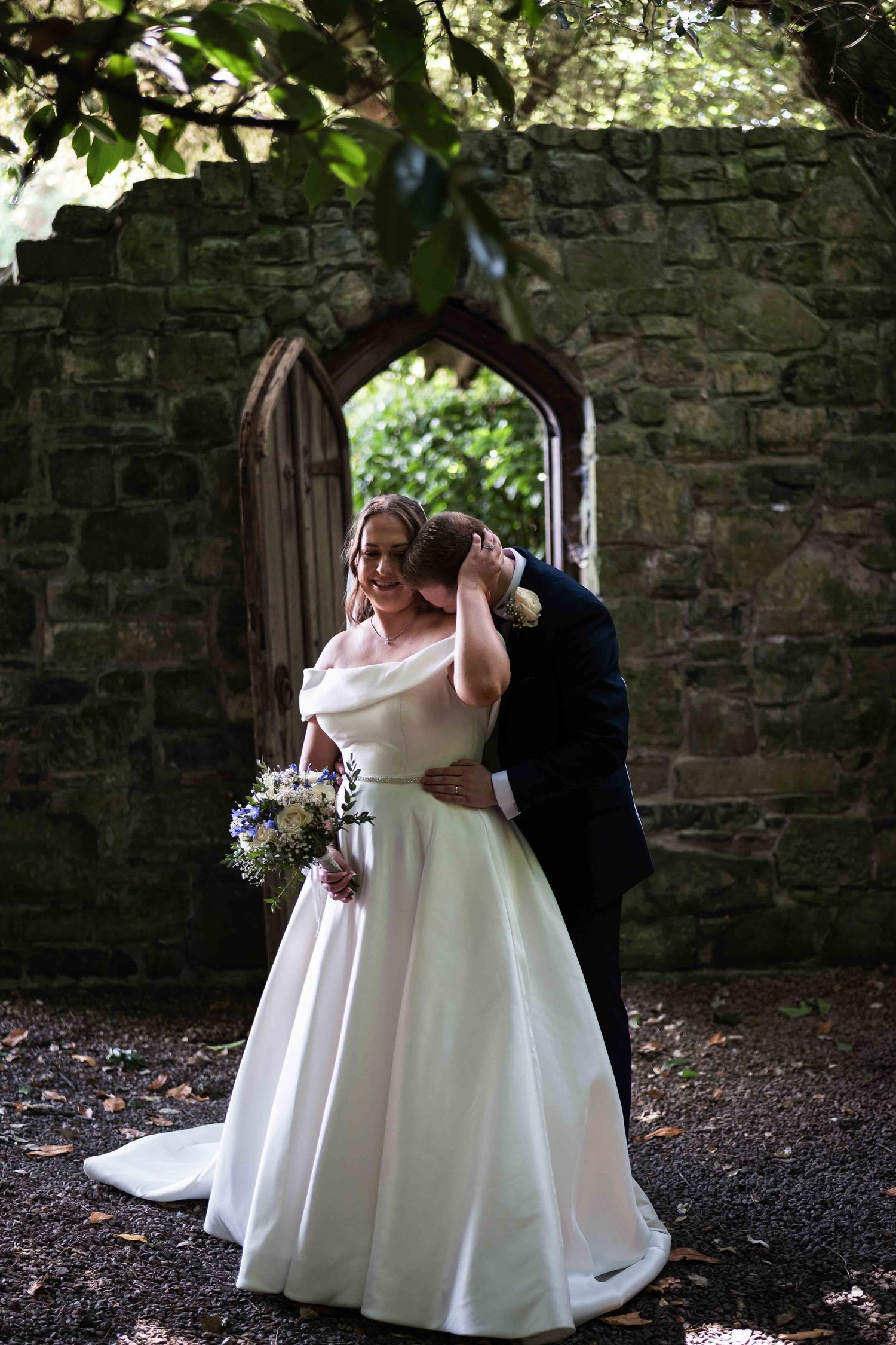 Bride in off-shoulder white gown holding bouquet embraced by groom in navy suit before stone archway. Dunboyne Castle
