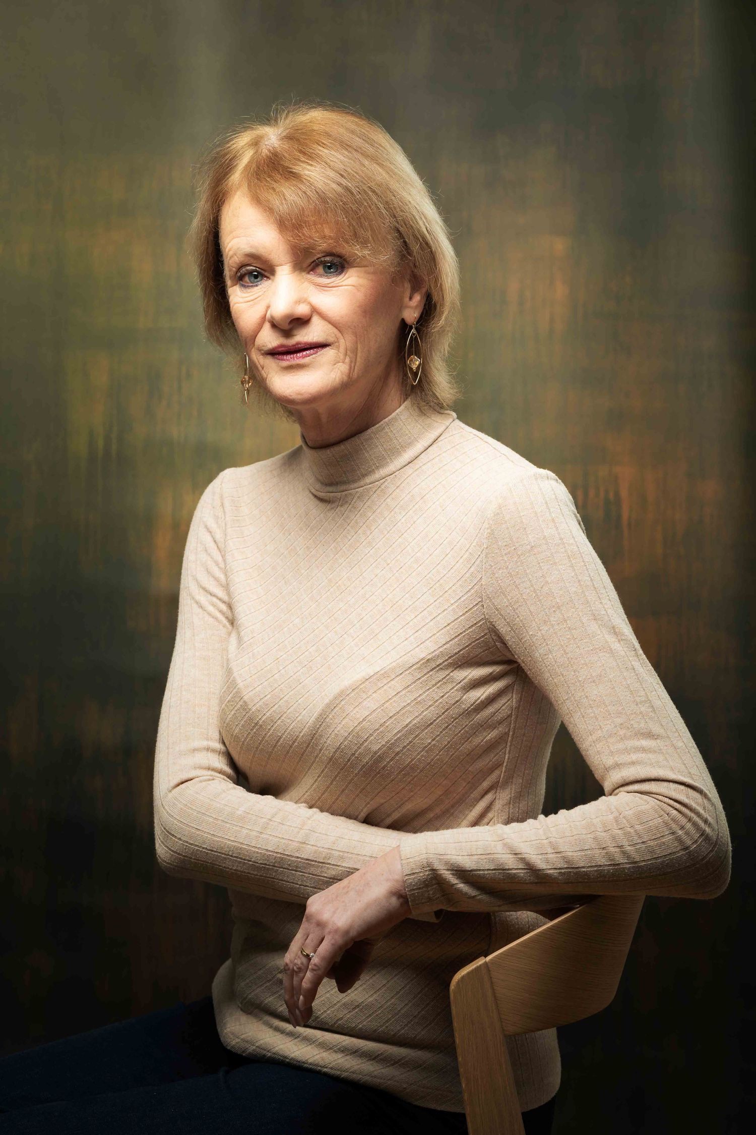 Middle-aged blonde woman in beige turtleneck sweater sitting on chair against dark textured background. Portrait Photography Ireland