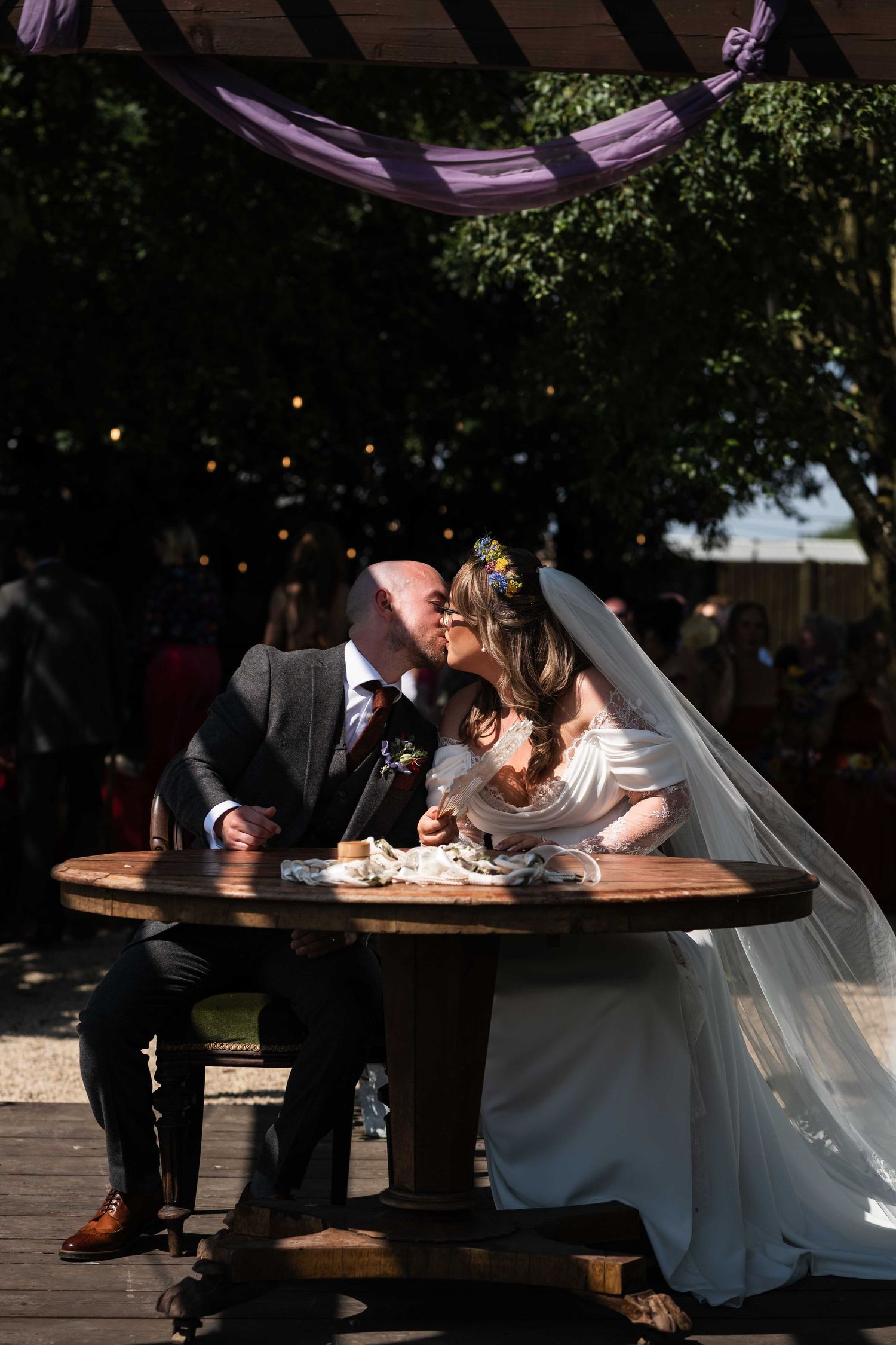 Bride and groom sharing a kiss at a wooden table during their outdoor wedding reception under a pergola. Mountain View, Kilkenny