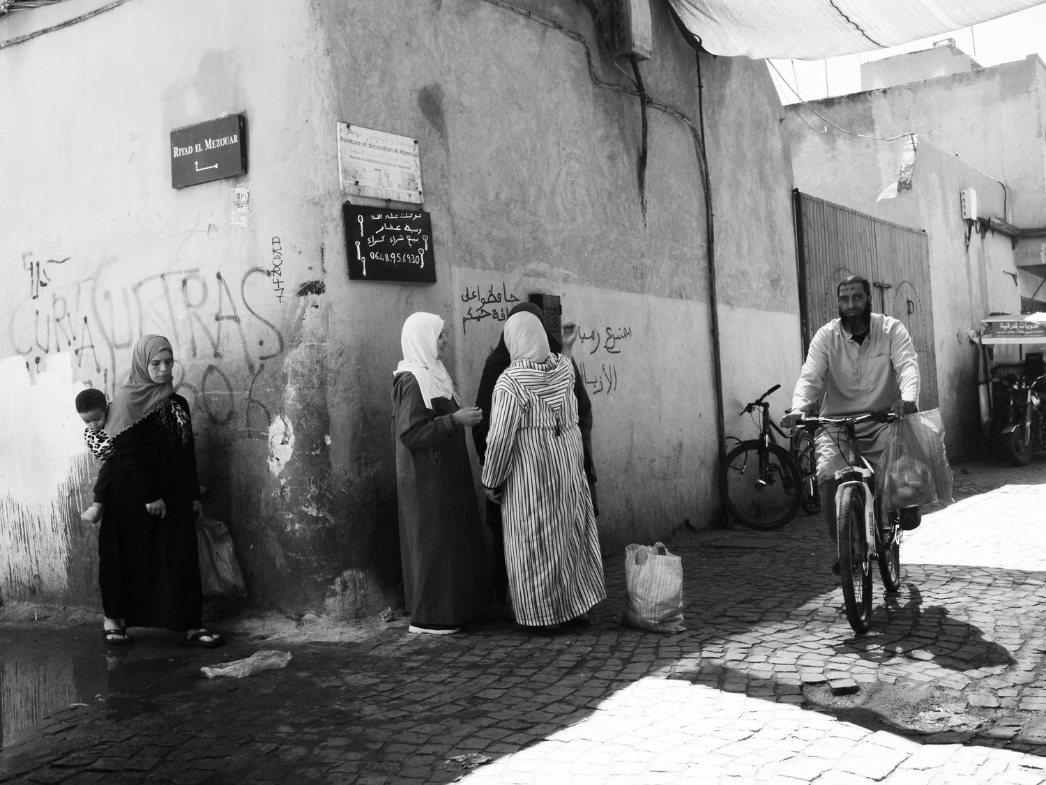 Black and white street scene with traditionally dressed women and a cyclist on cobblestone streets in Morocco.