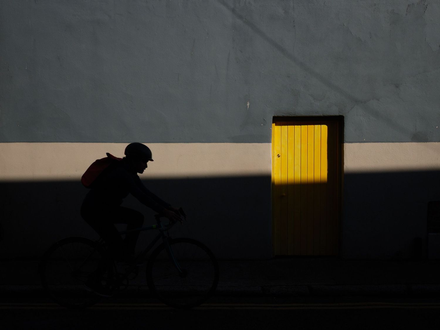 Silhouette of a cyclist riding past a glowing yellow door against a grey wall at golden hour.