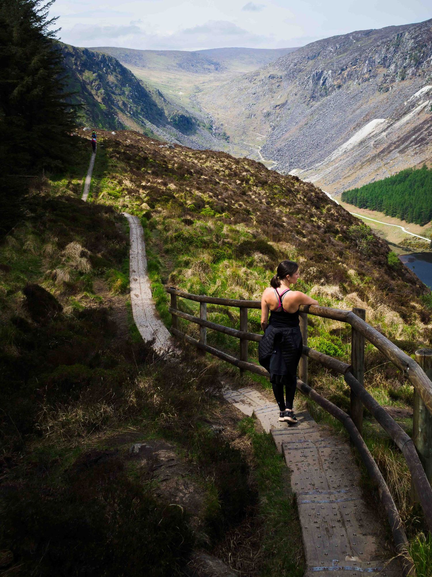 Woman hiking on wooden boardwalk trail with scenic Irish mountain valley landscape in background.