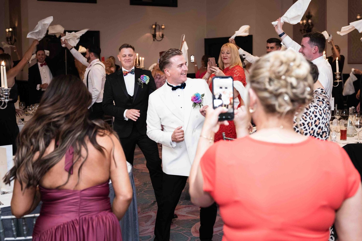 Groom in white tuxedo celebrates with guests waving napkins during lively wedding reception entrance. Queer wedding. Clontarf Castle