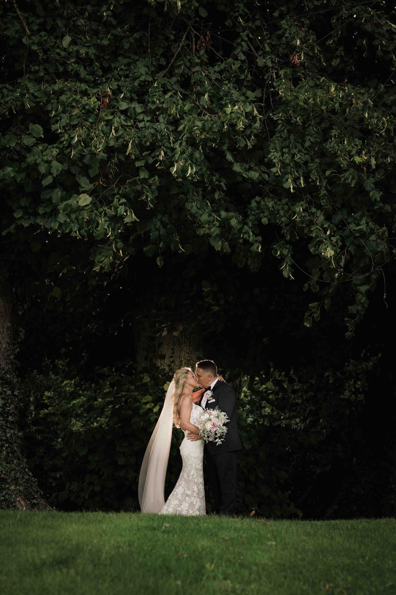 Bride and groom share romantic kiss under large tree at night holding white wedding bouquet. Dunboyne Castle