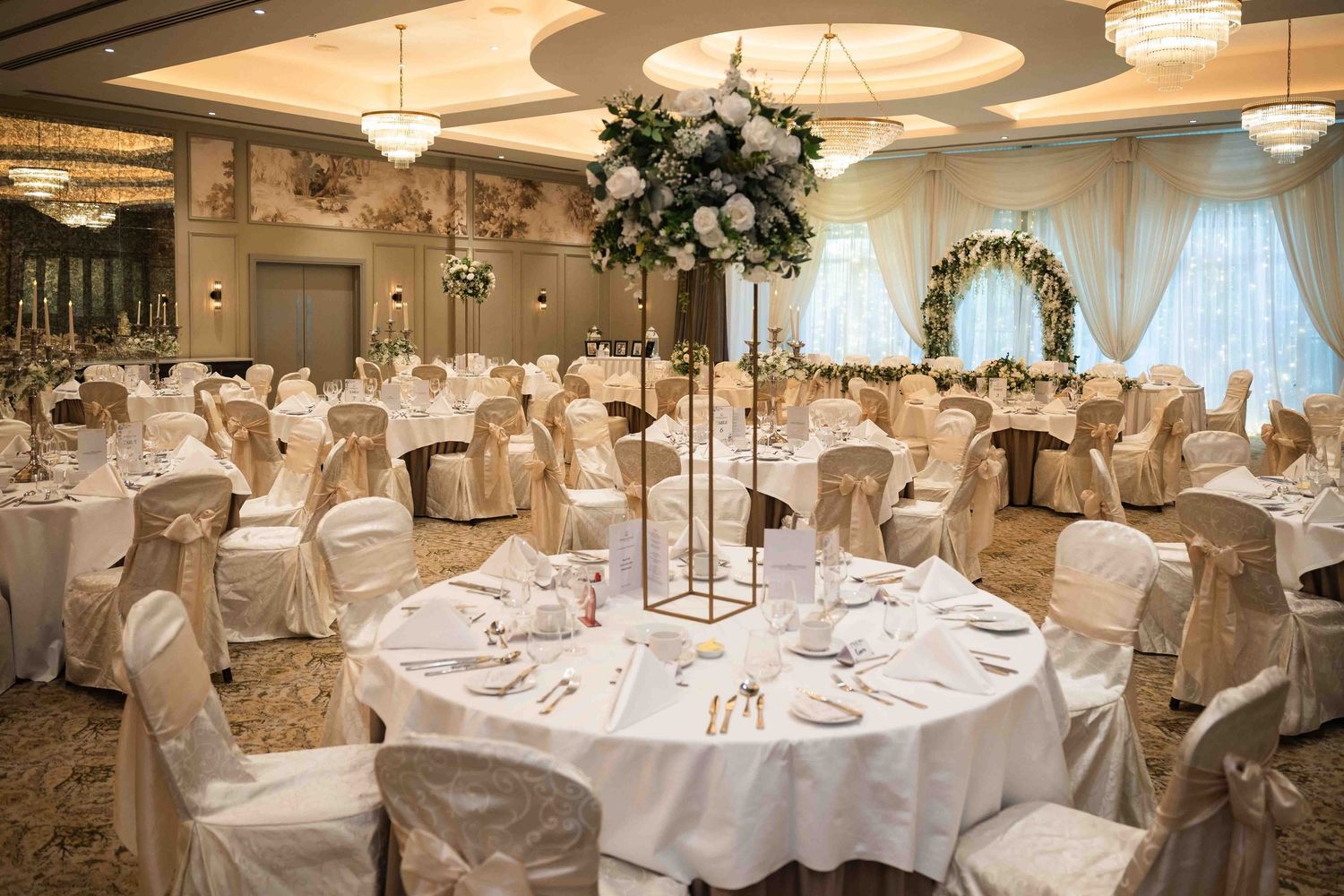 Elegant wedding reception ballroom with white chair covers, round tables, tall floral centerpieces, and soft ivory decor. Dunboyne Castle