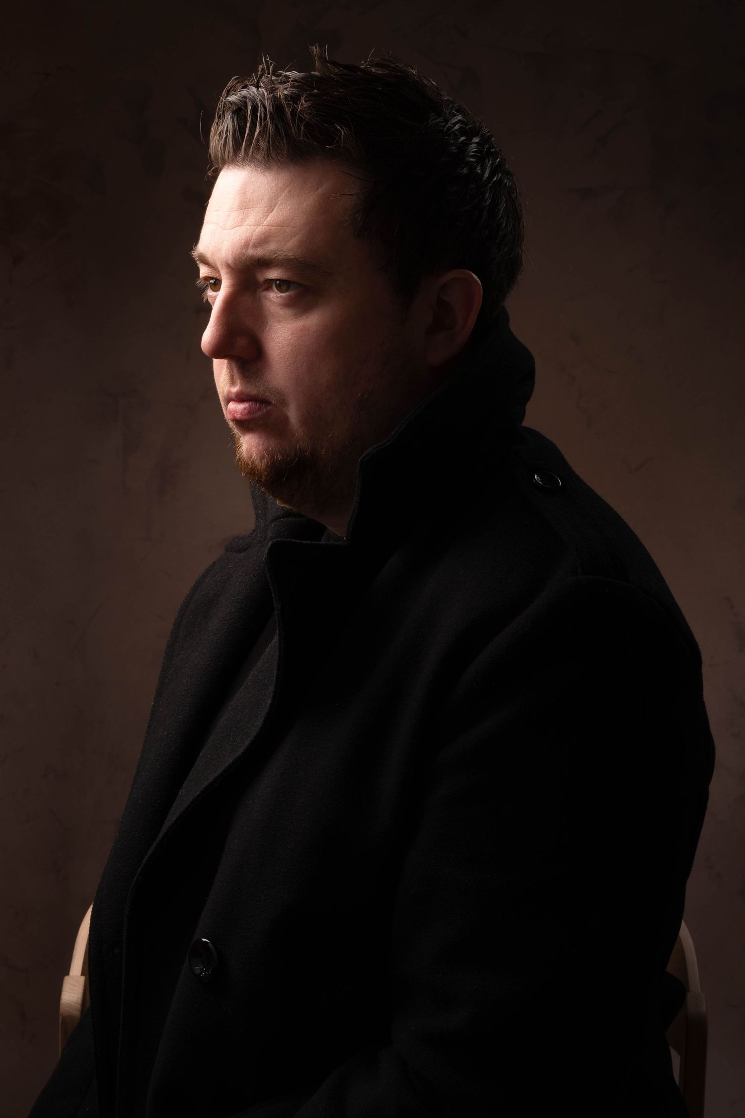 Dark-haired bearded man in black coat photographed in dramatic side profile against dark background. Portrait Photography Ireland