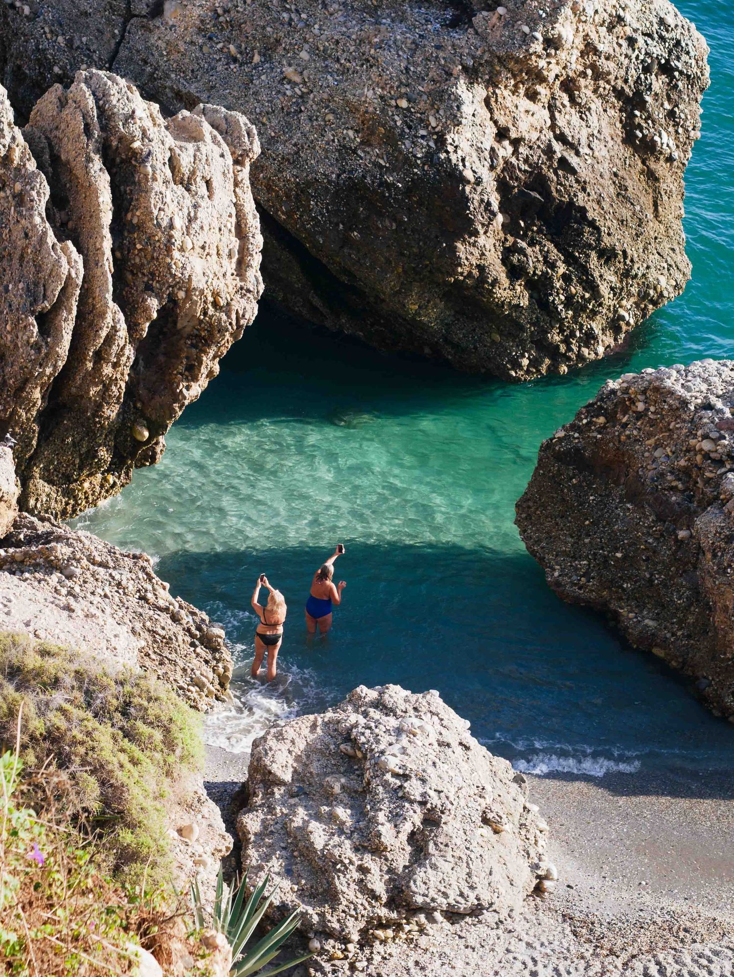 Two swimmers stand at the edge of a secluded turquoise cove surrounded by dramatic rocky cliffs.