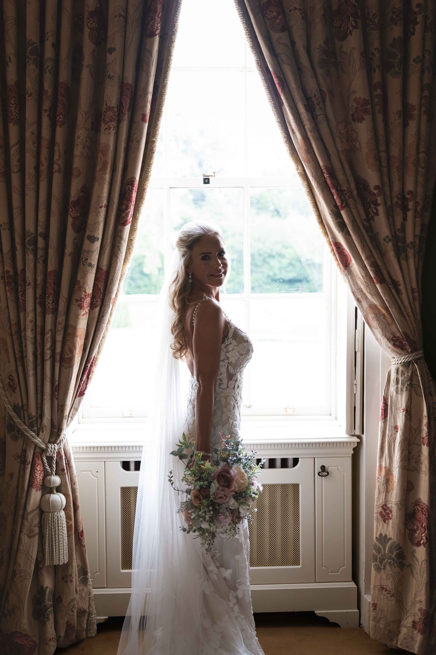 Bride in lace wedding dress holding bouquet stands by floral curtained window in elegant room. Dunboyne Castle