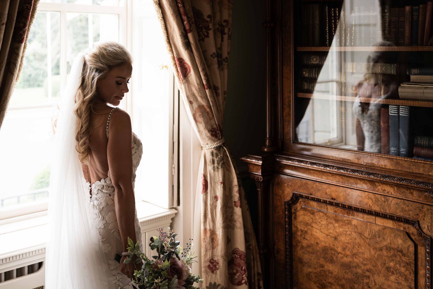 Bride in lace wedding dress holding bouquet stands by ornate window with floral curtains in elegant wood-paneled room. Dunboyne Castle