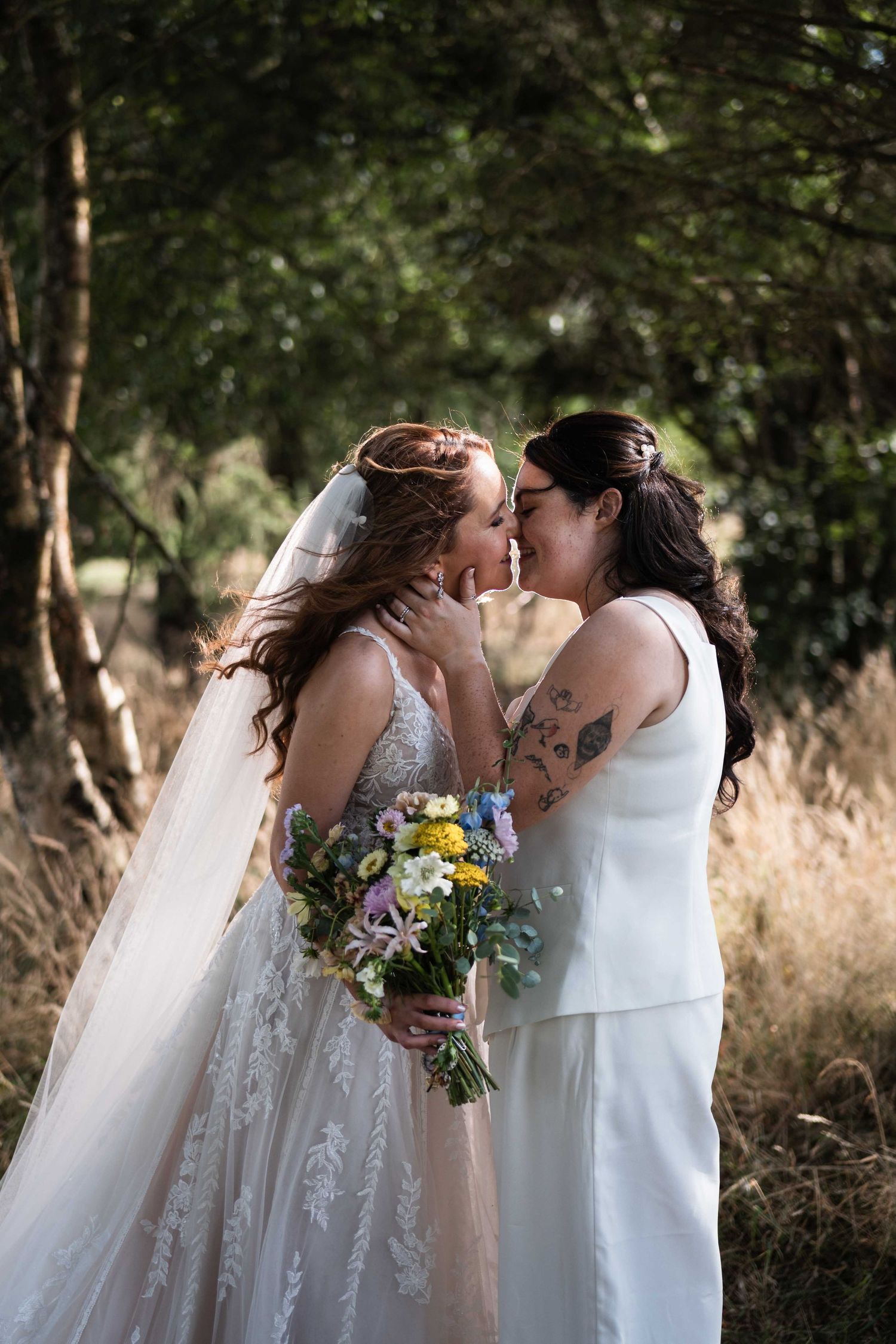 Two brides sharing a kiss outdoors, one holding a wildflower bouquet in natural light. Irish wedding photography. Mountain View, Kilkenny