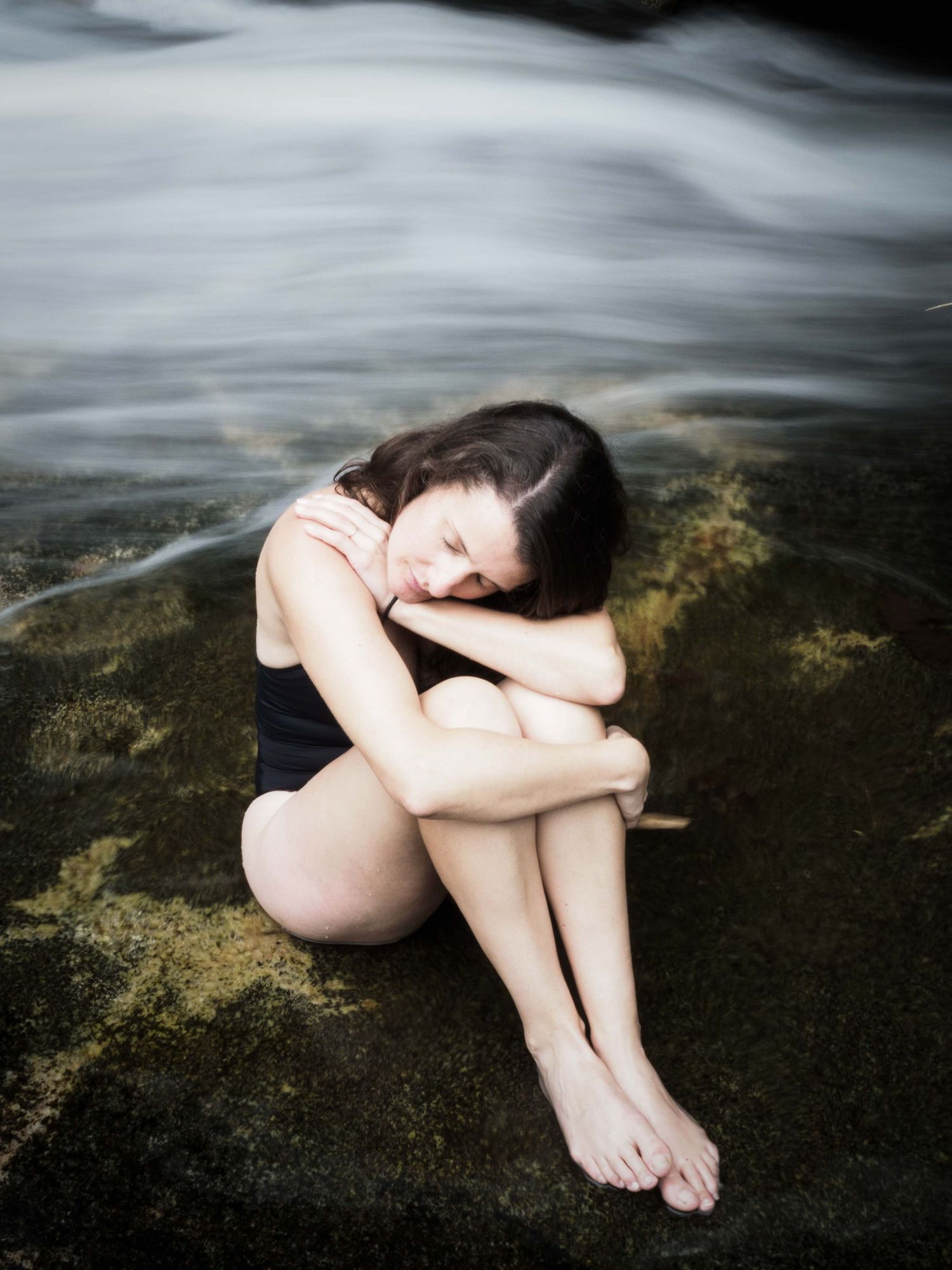 Young woman in black swimsuit sitting curled up on mossy rocks beside flowing water. Portrait Photography Ireland