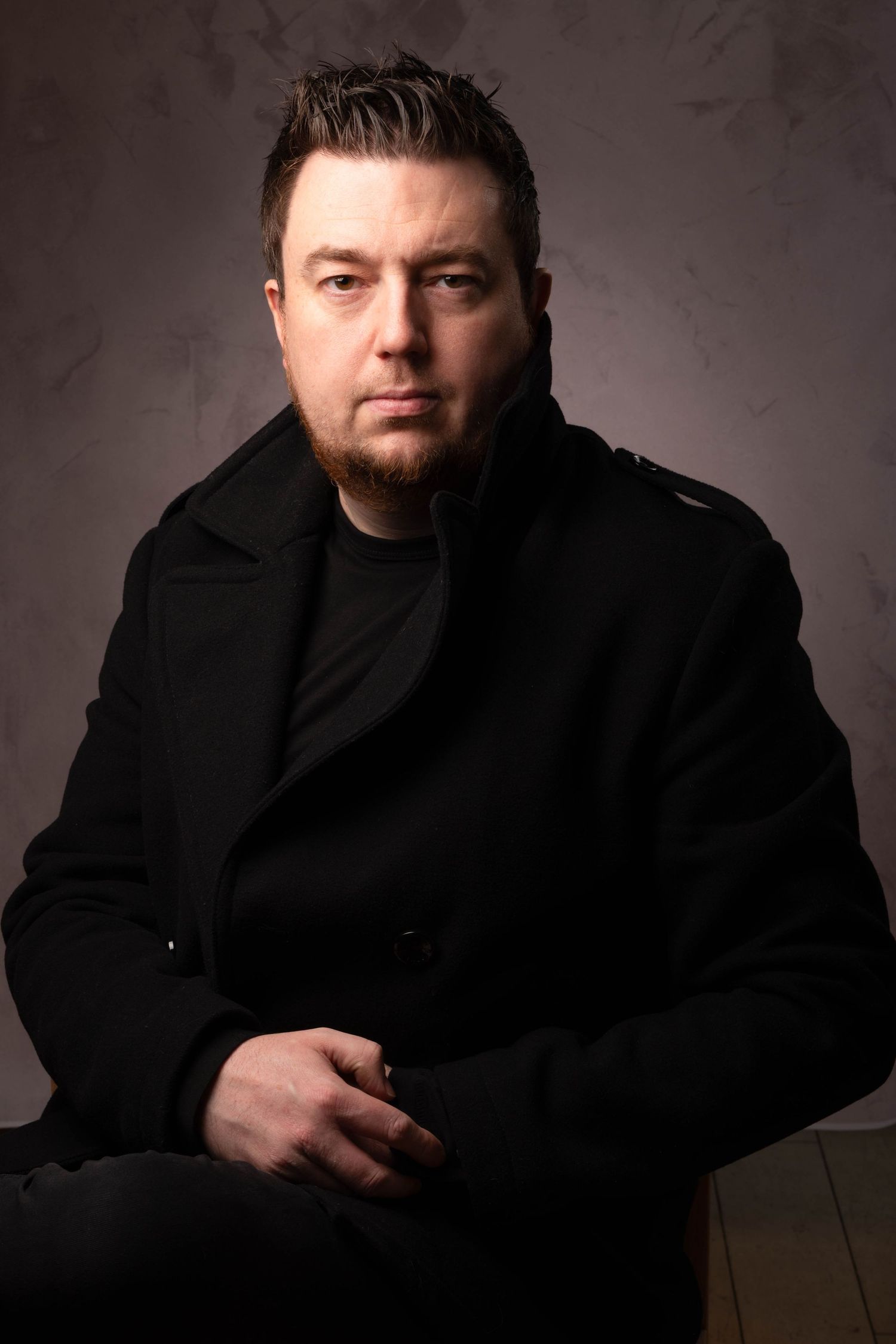 Bearded man in black coat sitting and looking at camera against gray textured background.  Portrait Photography Ireland