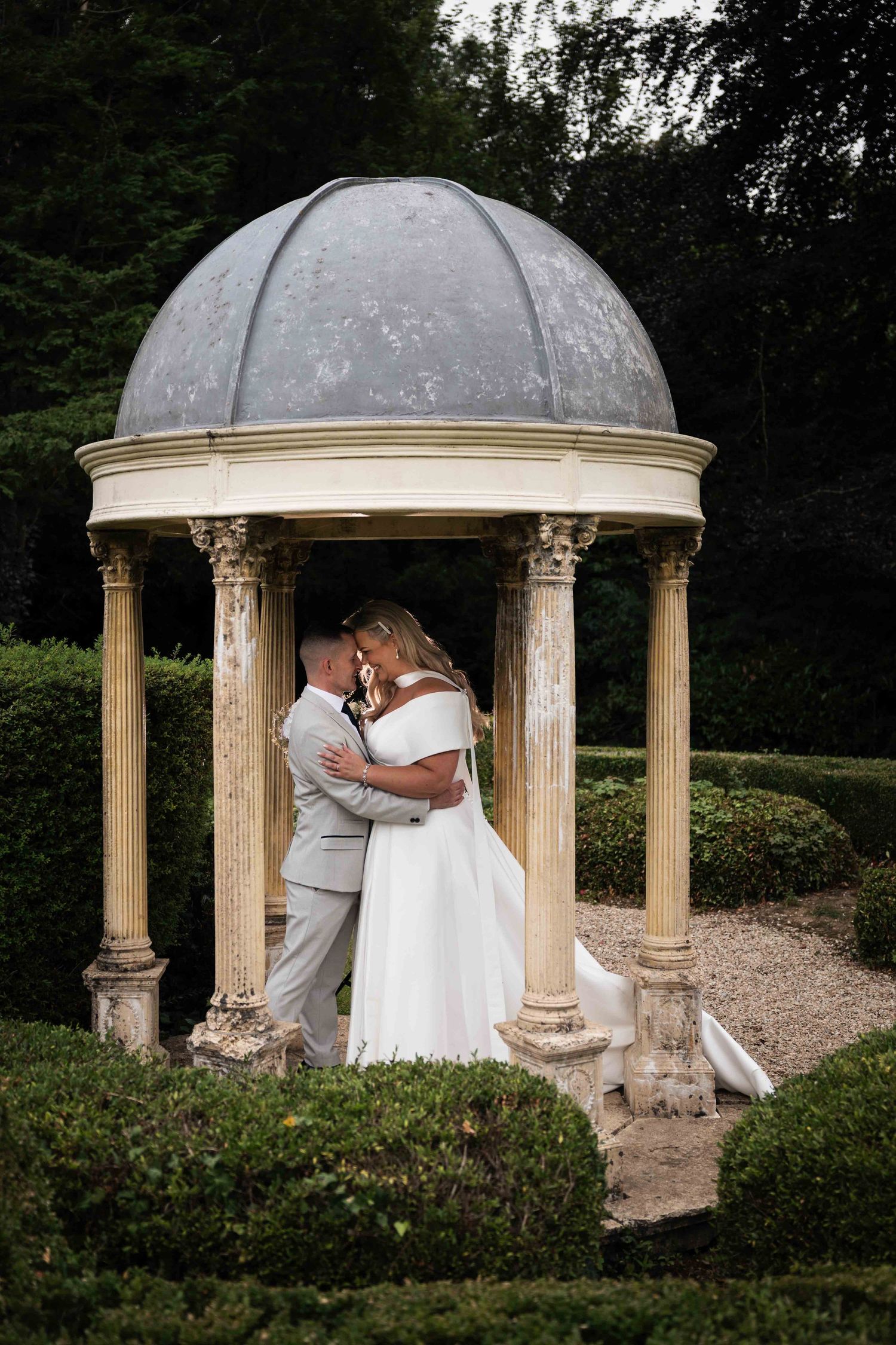 Bride and groom embracing under an elegant stone gazebo with domed roof in a formal garden setting. Dunboyne Castle