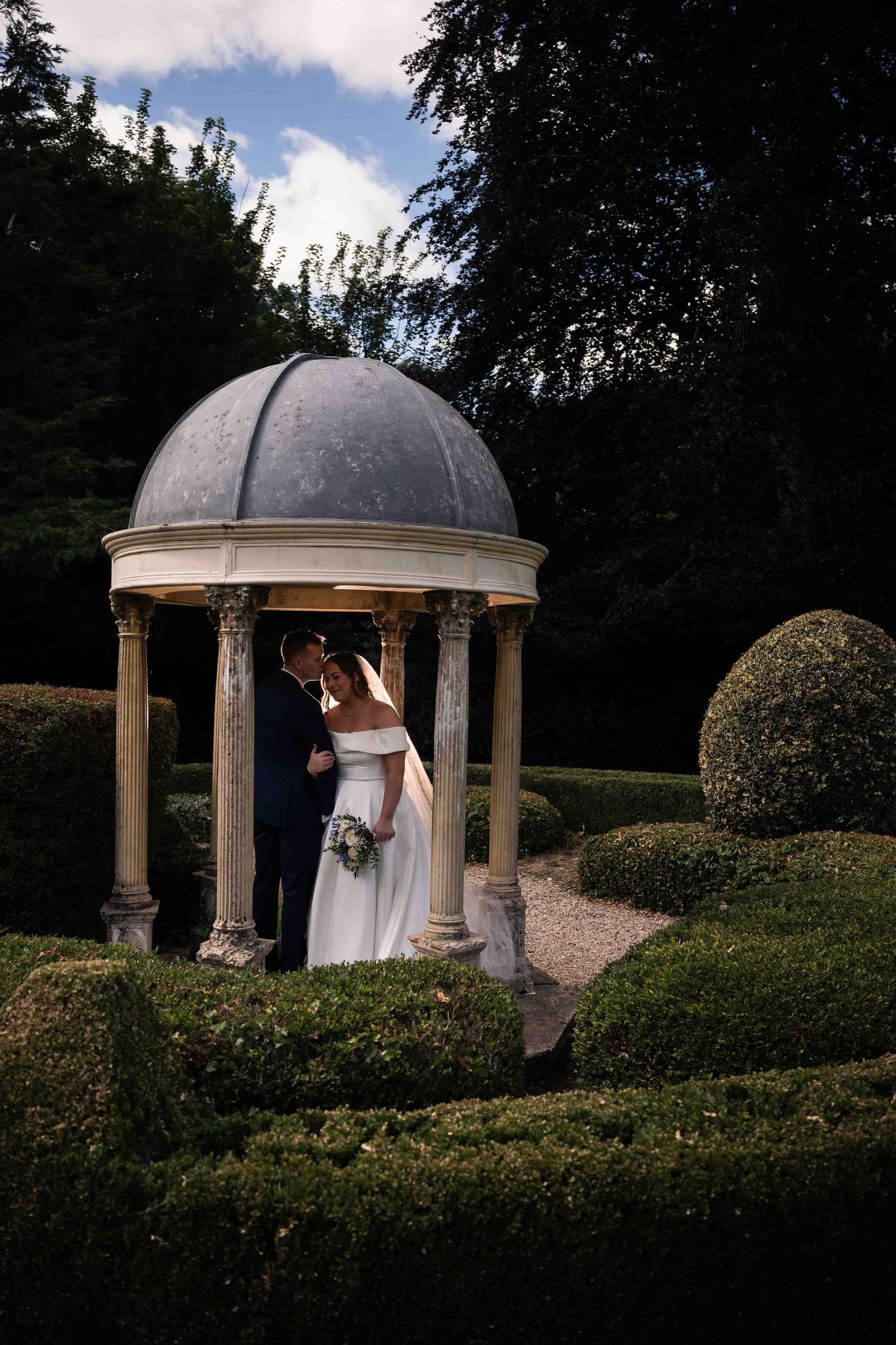Bride and groom share a kiss under a stone domed gazebo surrounded by manicured hedges. Irish wedding photography. Dunboyne Castle