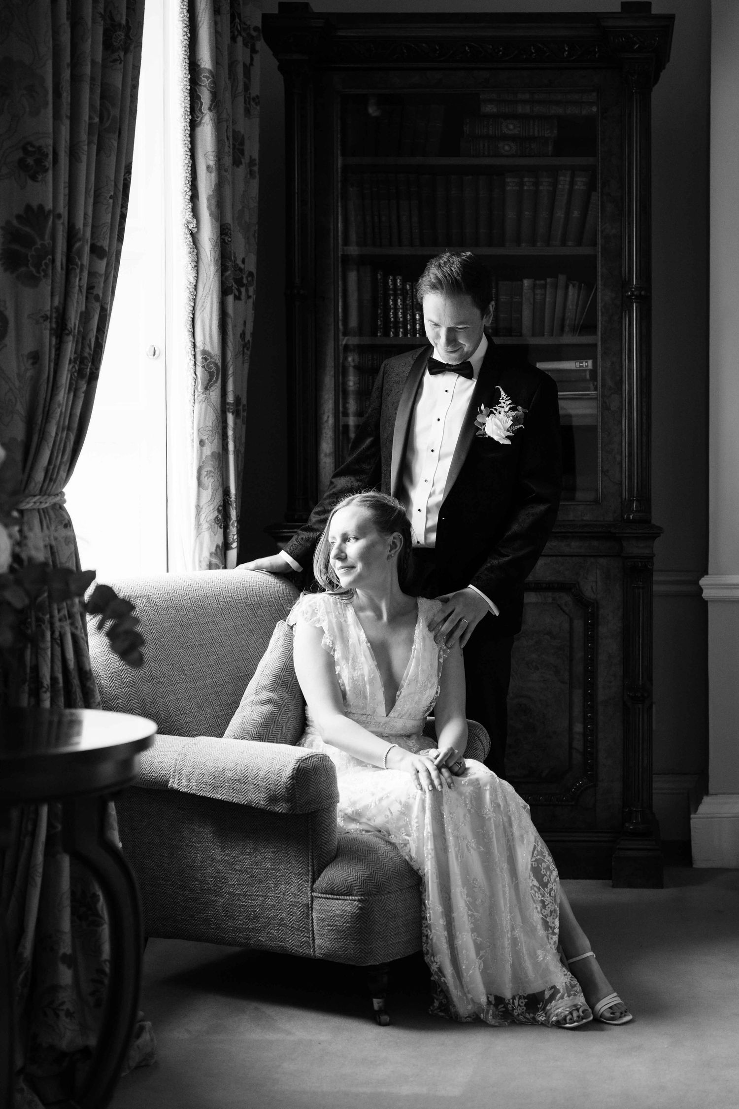 Black and white wedding photo of bride in lace gown seated in armchair with groom in tuxedo standing beside her. Dunboyne Castle