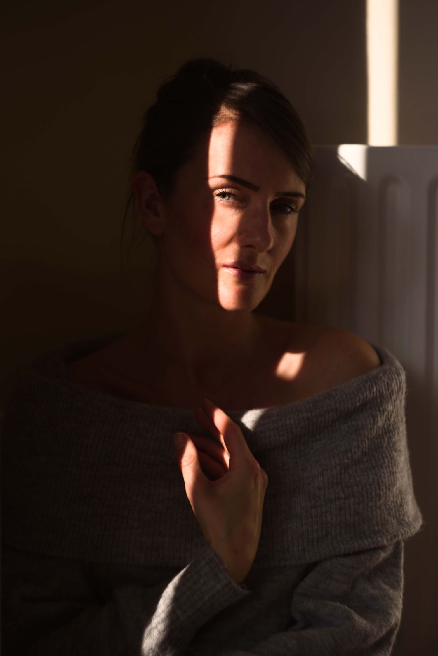 Woman in an off-shoulder grey sweater posed in dramatic chiaroscuro lighting with sunlight on her shoulder. Portrait Photography Ireland
