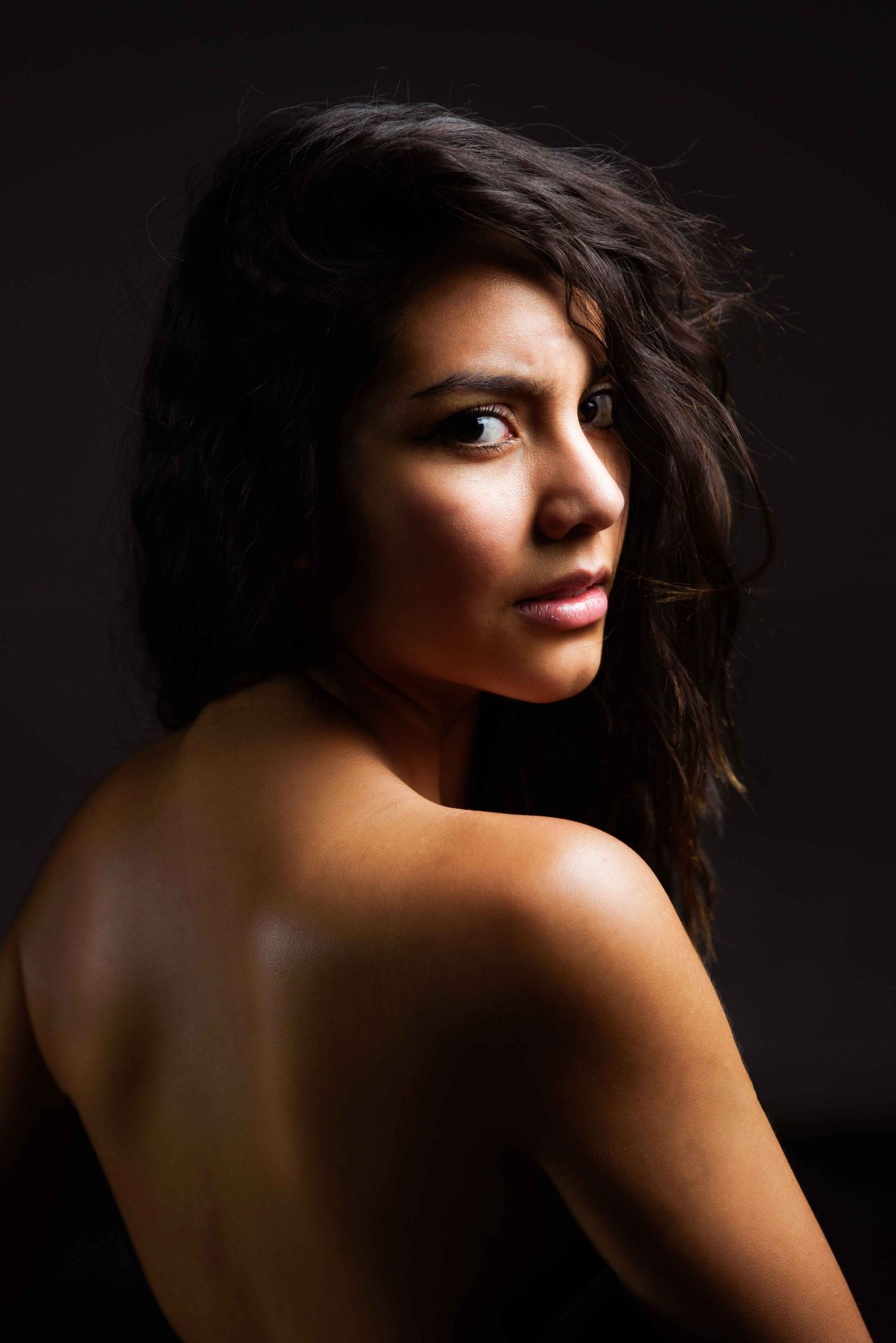 Brunette woman with bare shoulders glancing back over her shoulder in dark studio lighting. Portrait Photography Ireland