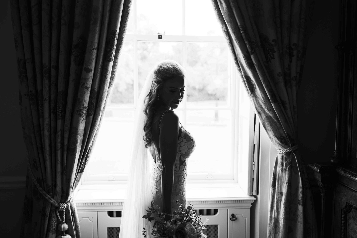 Black and white silhouette of a bride in veil standing by a curtained window holding flowers. Dunboyne Castle